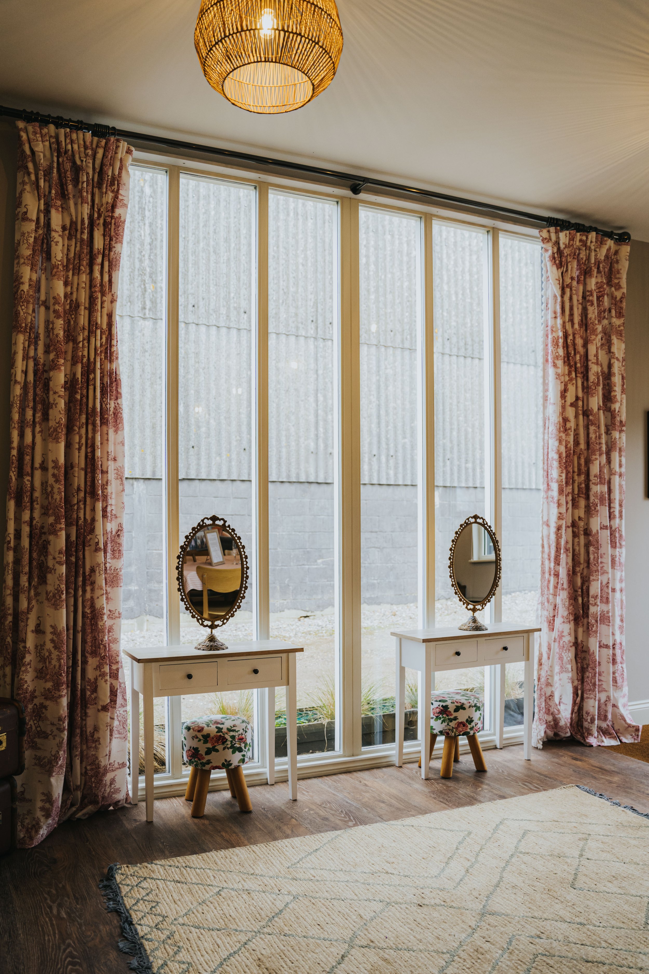 A room with large window panes, floral curtains, two white vanity tables with mirrors, small upholstered stools, and a beige rug on wooden floor.