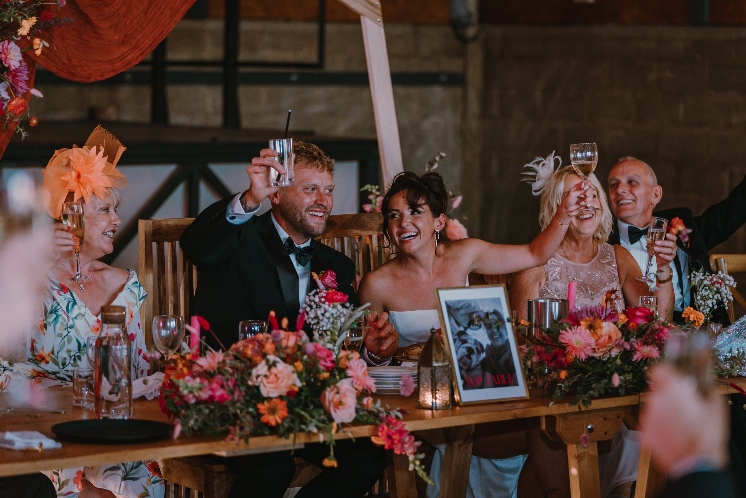 Wedding reception with guests raising glasses in toast, seated at a decorated table with floral arrangements, a framed photo, and candles.