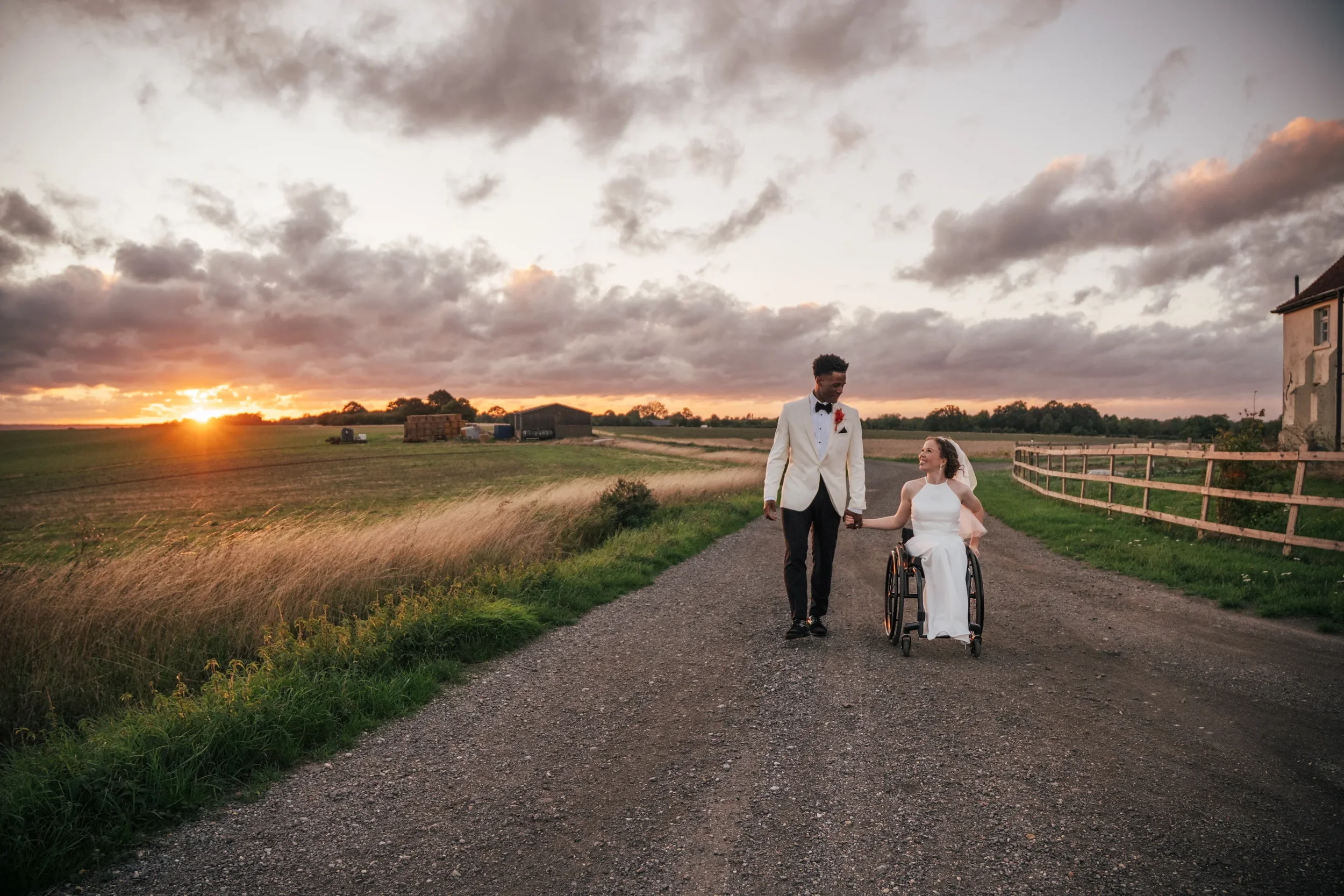 A couple dressed in wedding attire walking hand in hand along a dirt road at sunset, with the woman in a wheelchair, in a rural setting with fields, a fence, and farm buildings in the background.