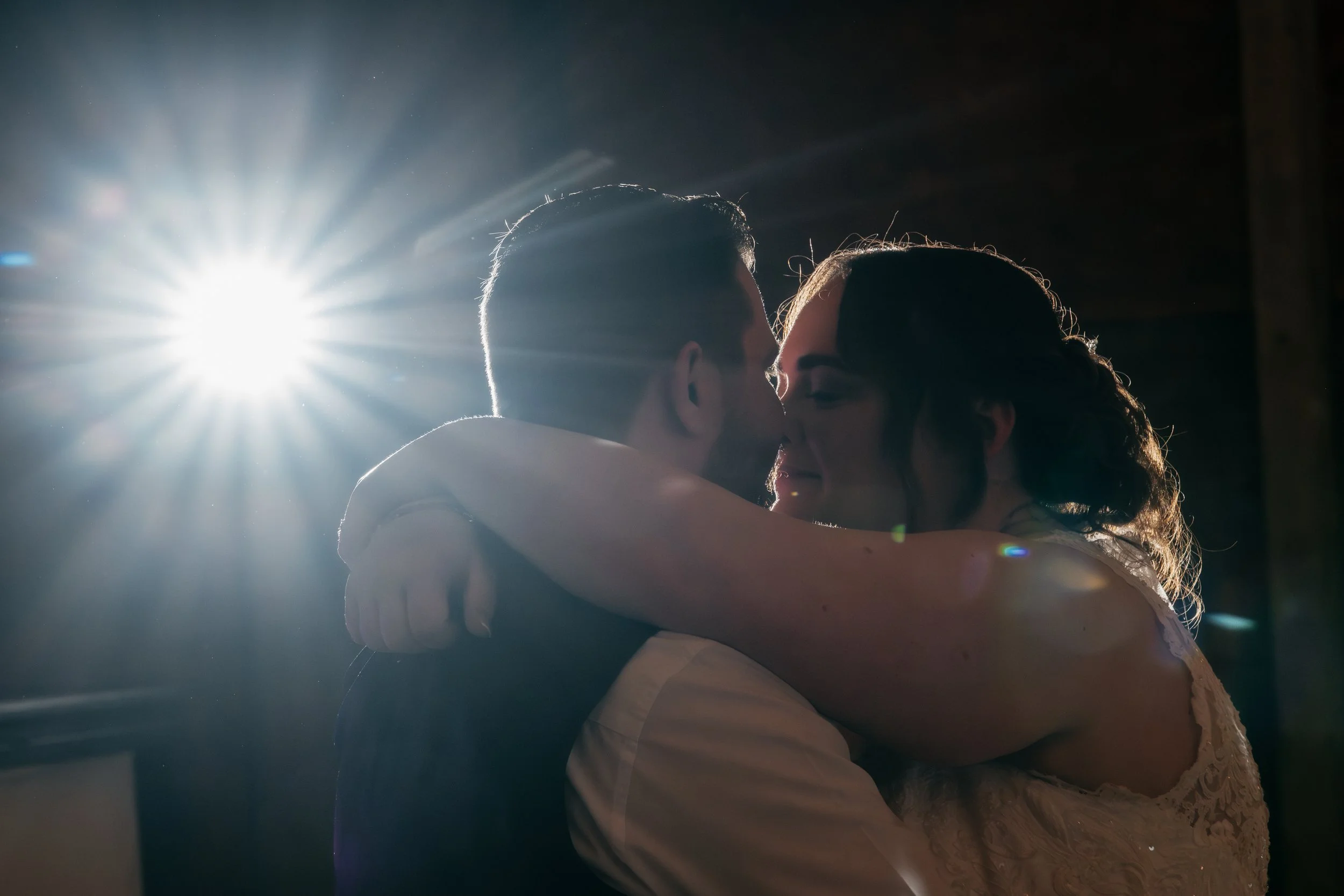 A couple in a close embrace, about to kiss or kiss, with bright backlighting creating a silhouette effect, during a special moment such as a wedding or celebration.