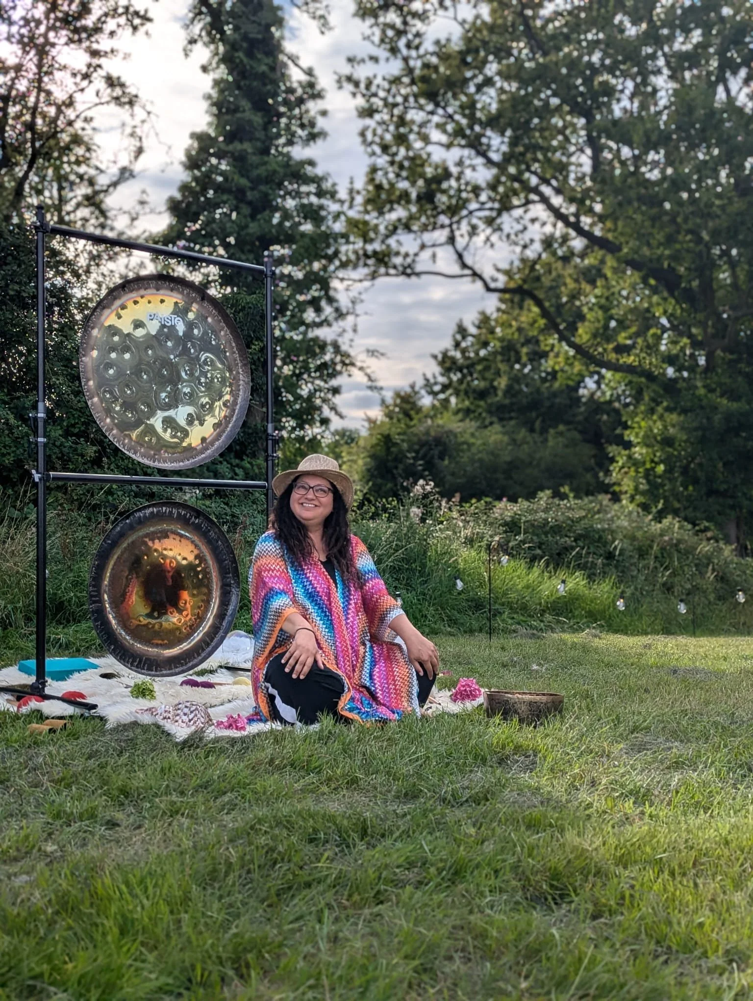 Woman sitting on a blanket outdoors in front of two large singing bowls, smiling, wearing glasses, a multicolored poncho, and a wide-brimmed hat, with green trees and a cloudy sky in the background.