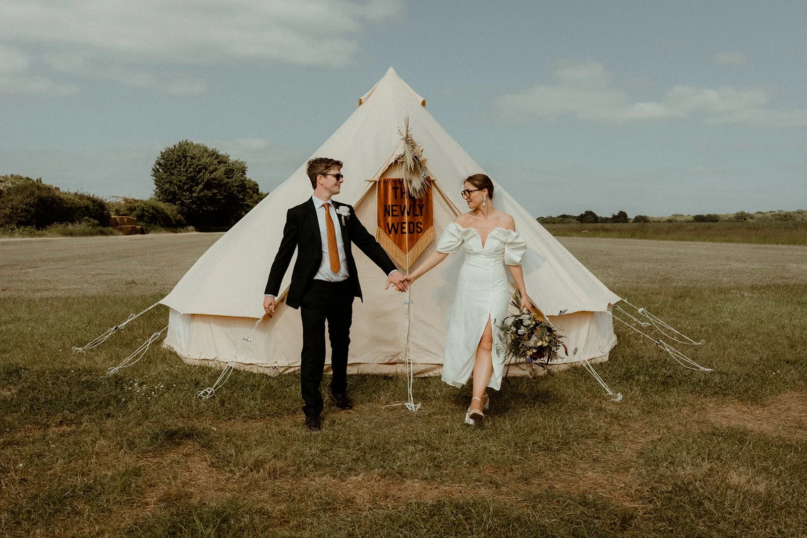 A newlywed couple holding hands in front of a white tent in an open field, with a sign inside the tent that reads 'The Newly Weds.' The groom wears a black suit with orange tie and the bride wears a white off-shoulder dress with high slit, holding a bouquet of flowers.