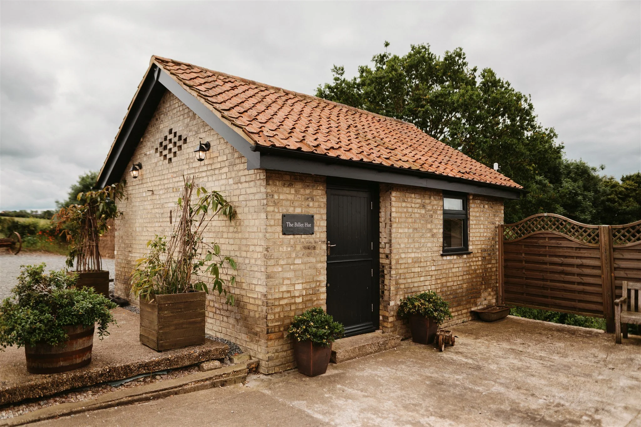 A small brick building with a red tiled gable roof, black door, and window. There are potted plants outside, a wooden fence on the right, and a cloudy sky overhead.