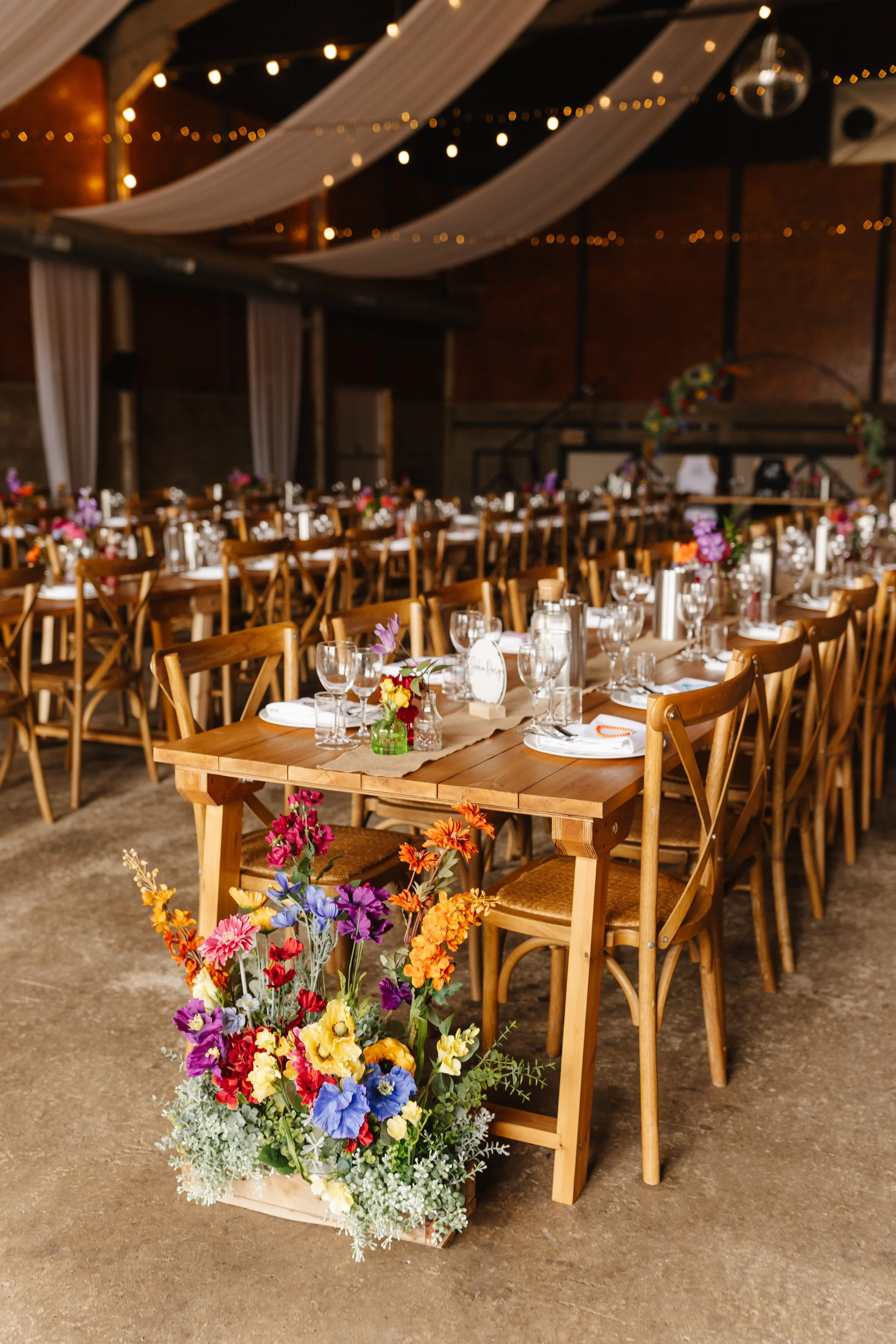 Decorated banquet hall with long wooden tables, floral arrangements, candle holders, and glassware, with draped ceiling and string lights for a celebration or wedding reception.