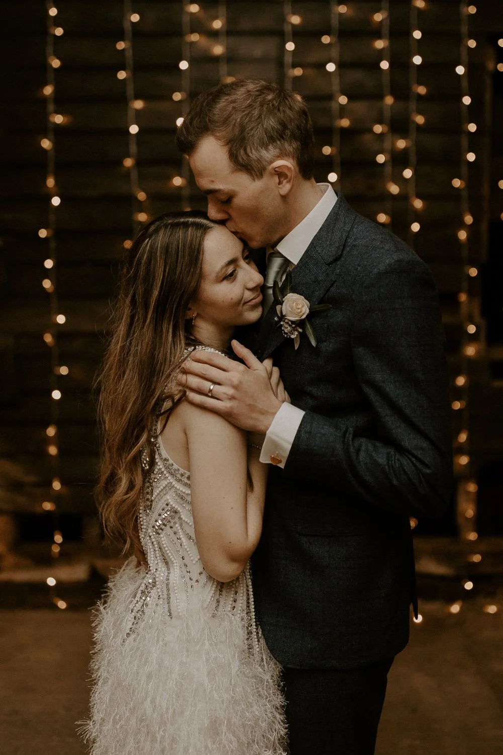 A couple on their wedding day sharing an intimate moment; the man is kissing the woman's forehead while she gently touches his chest, in a warmly lit setting with string lights in the background.