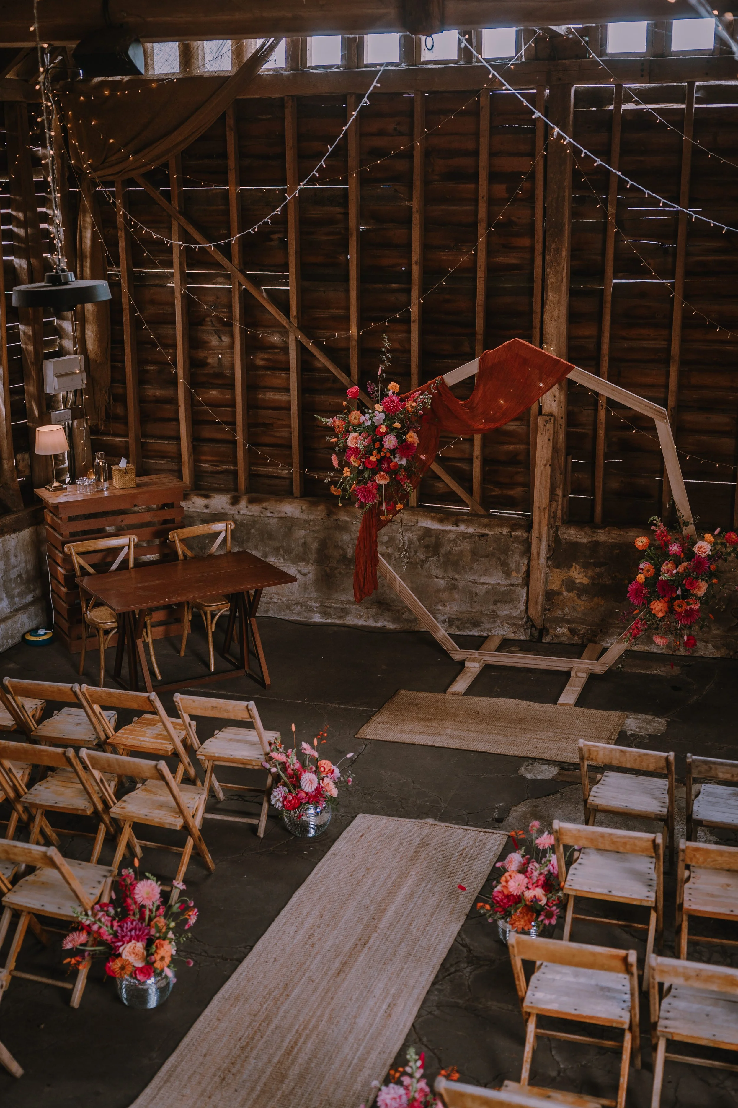 Inside a rustic barn decorated for a wedding ceremoy with a wooden floral arch and rows of wooden chairs with pink and orange flowers in vases, fairy lights strung across the ceiling.