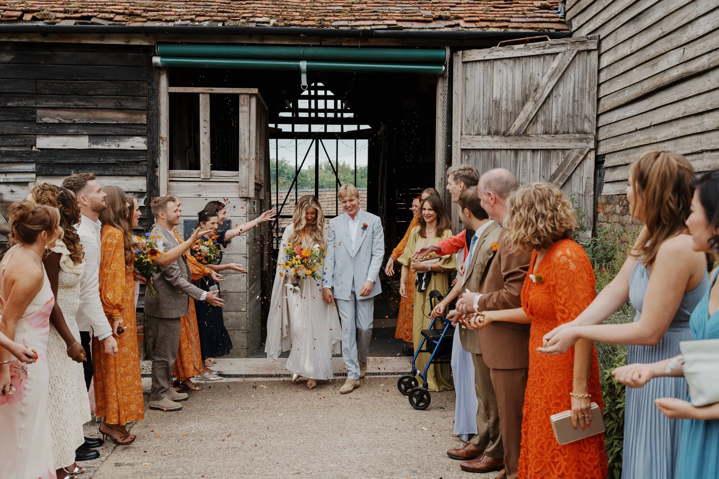 A newlywed couple walking out of a barn with friends and family celebrating their wedding, throwing confetti.