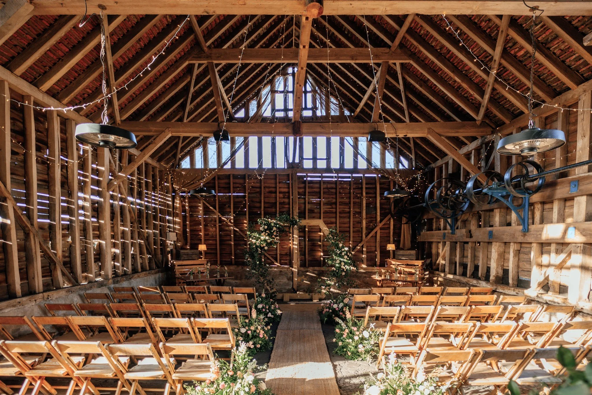 A rustic barn wedding setup with wooden chairs arranged in rows on either side of a central aisle lined with bouquets. The barn has wooden walls and a high, exposed beam ceiling decorated with string lights, creating a warm and festive atmosphere.