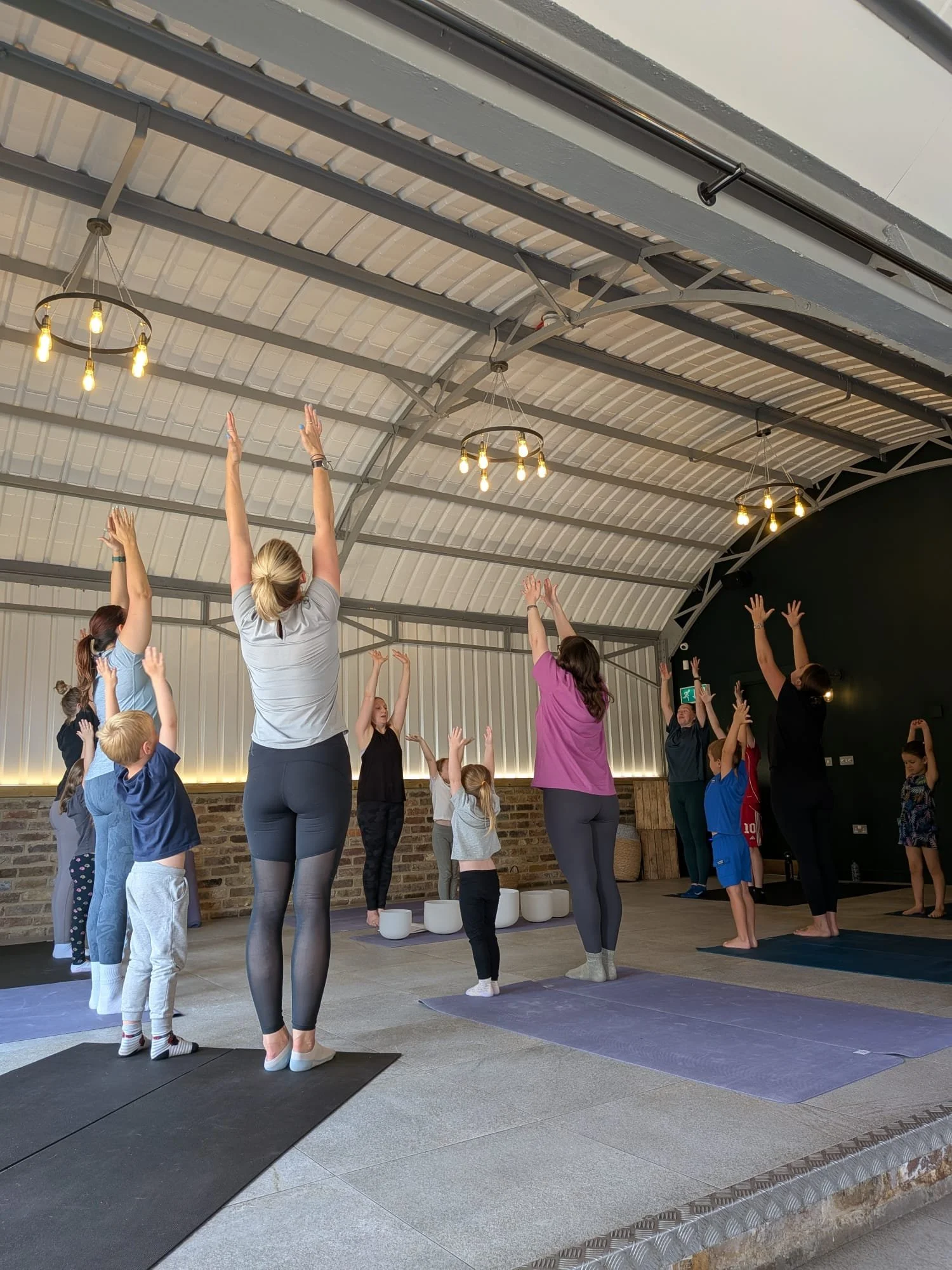 Group of women and children doing yoga in a spacious indoor studio with high arched ceiling and hanging lights.