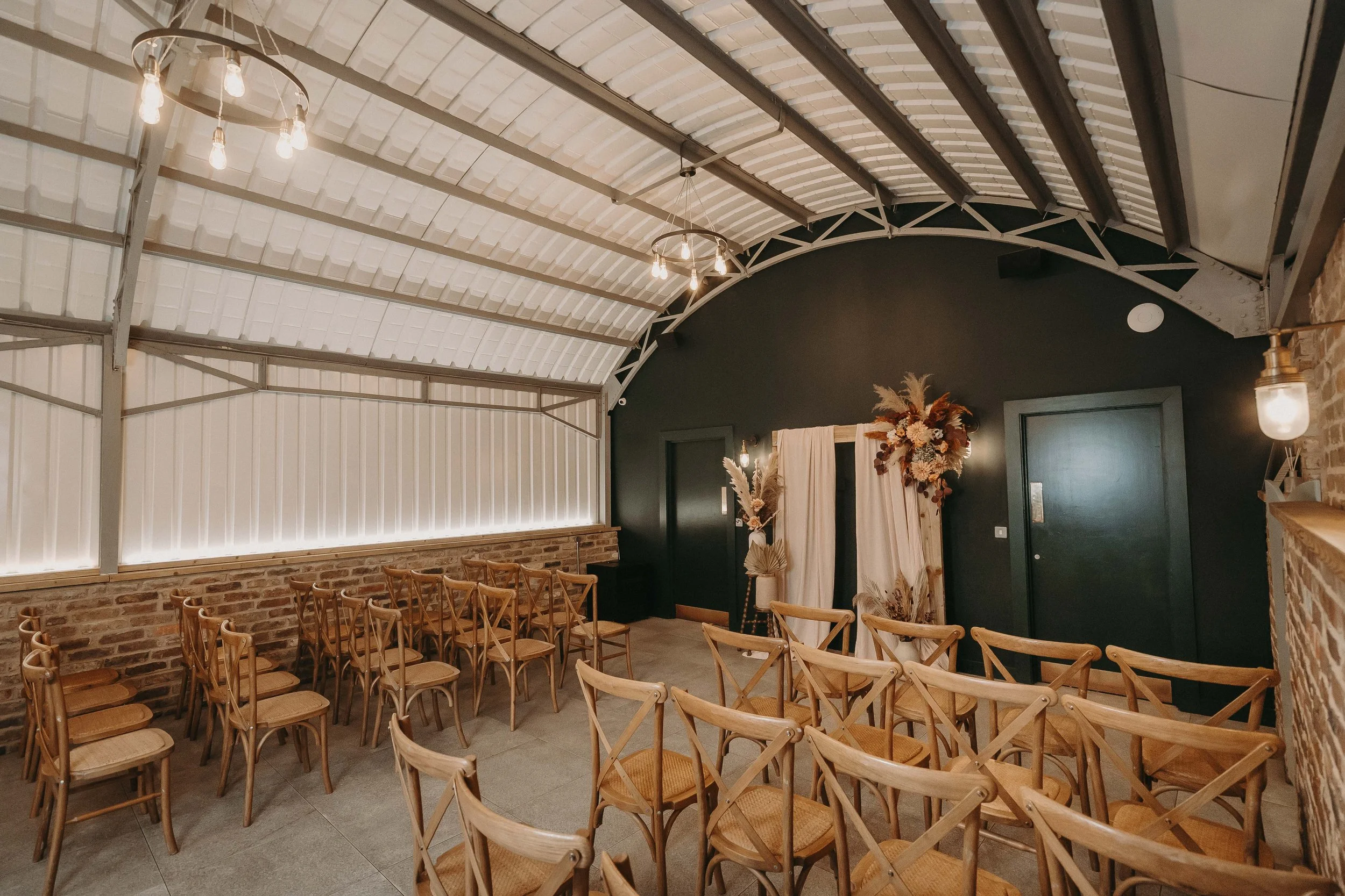 An indoor event space with rows of wooden chairs arranged facing a black accent wall, decorated with a floral arrangement and a curtain. Features include brick and brick accent walls, exposed ceiling beams, and hanging light fixtures.