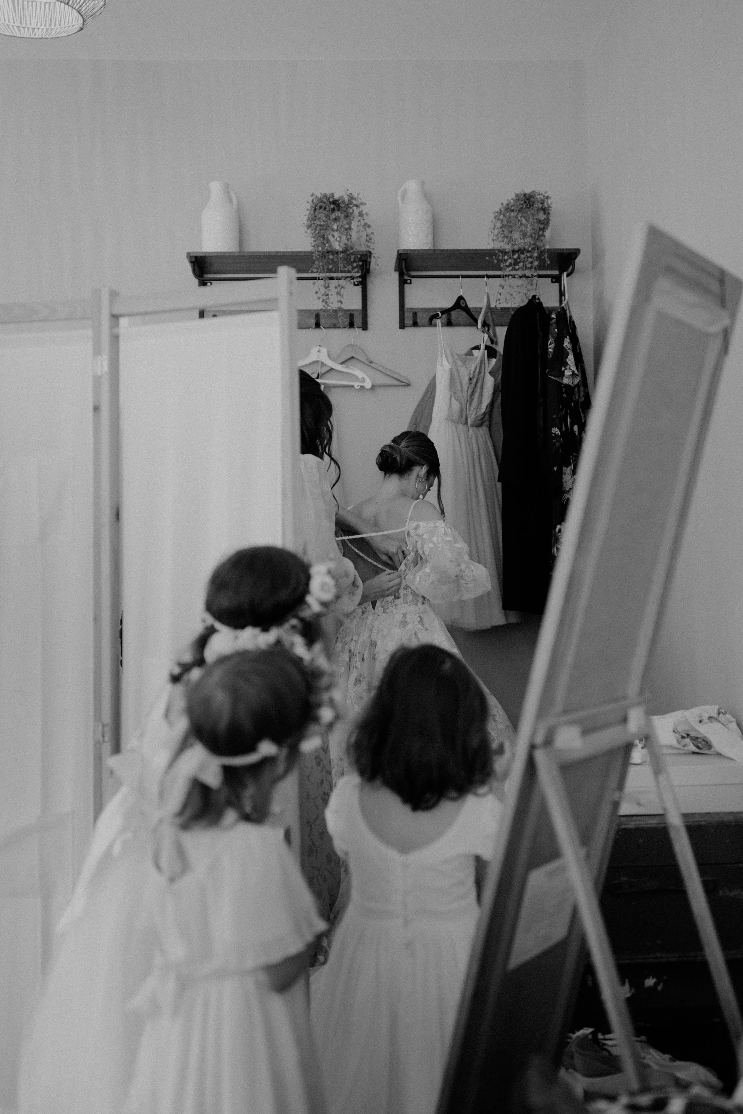 Women and children preparing dresses in a room, with dresses hanging on the wall, and a mirror reflecting a woman adjusting her dress.