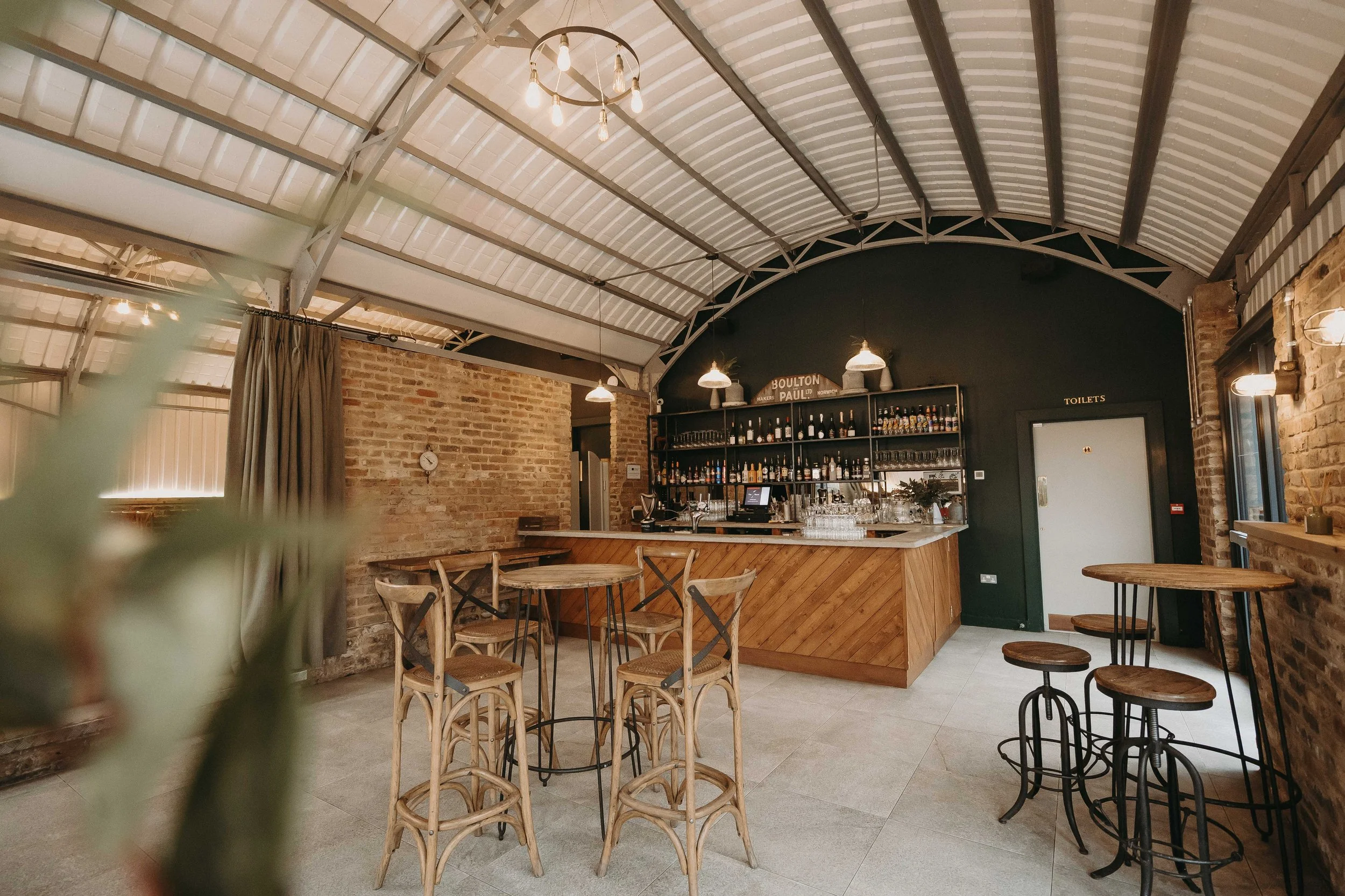 Interior of a modern bar with a curved metal roof, wooden furniture, a dark green wall, and a fully stocked bar counter.