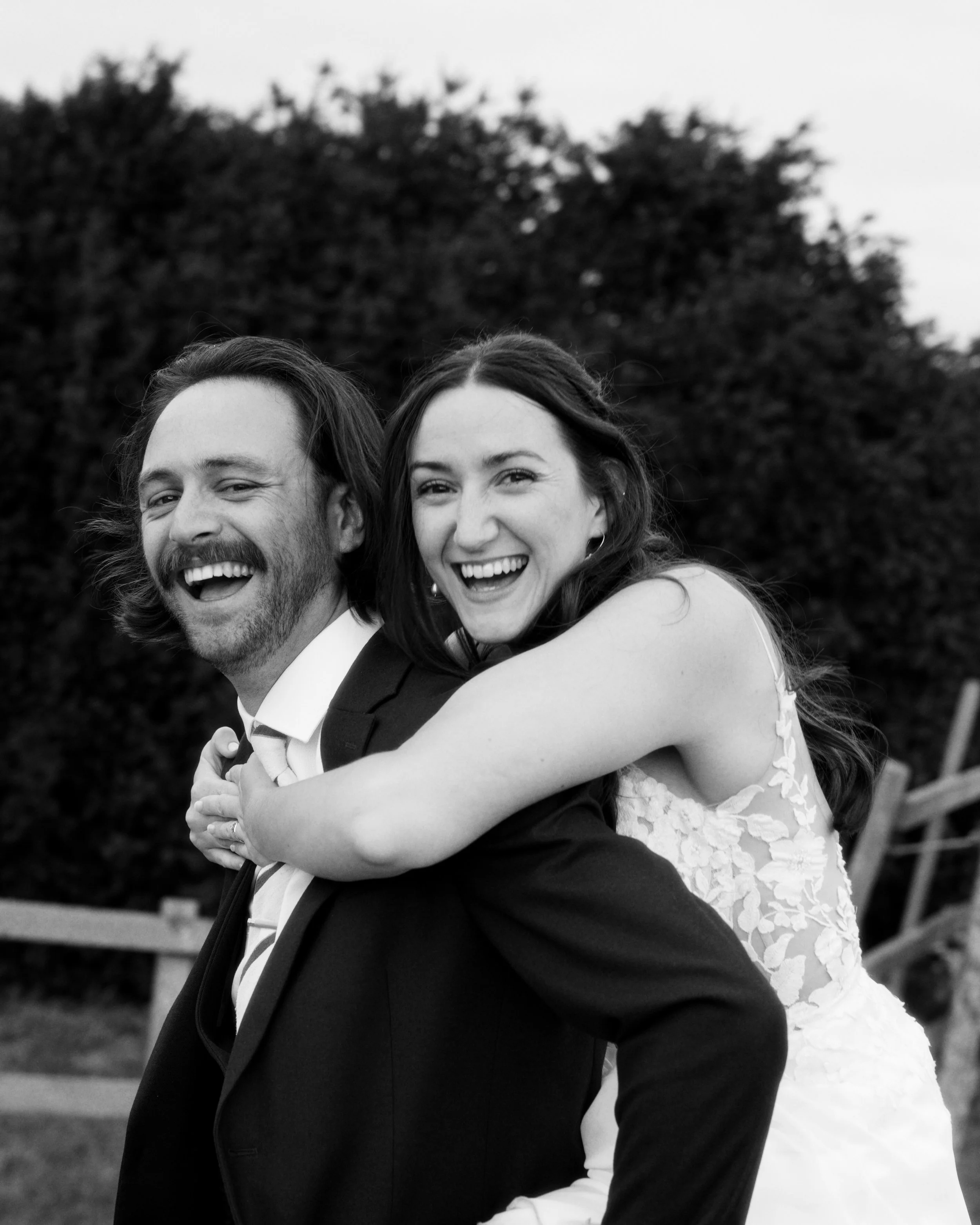Black and white photo of a happy man in a suit with a woman in a wedding dress on his back, smiling with their faces close together outdoors in front of trees.
