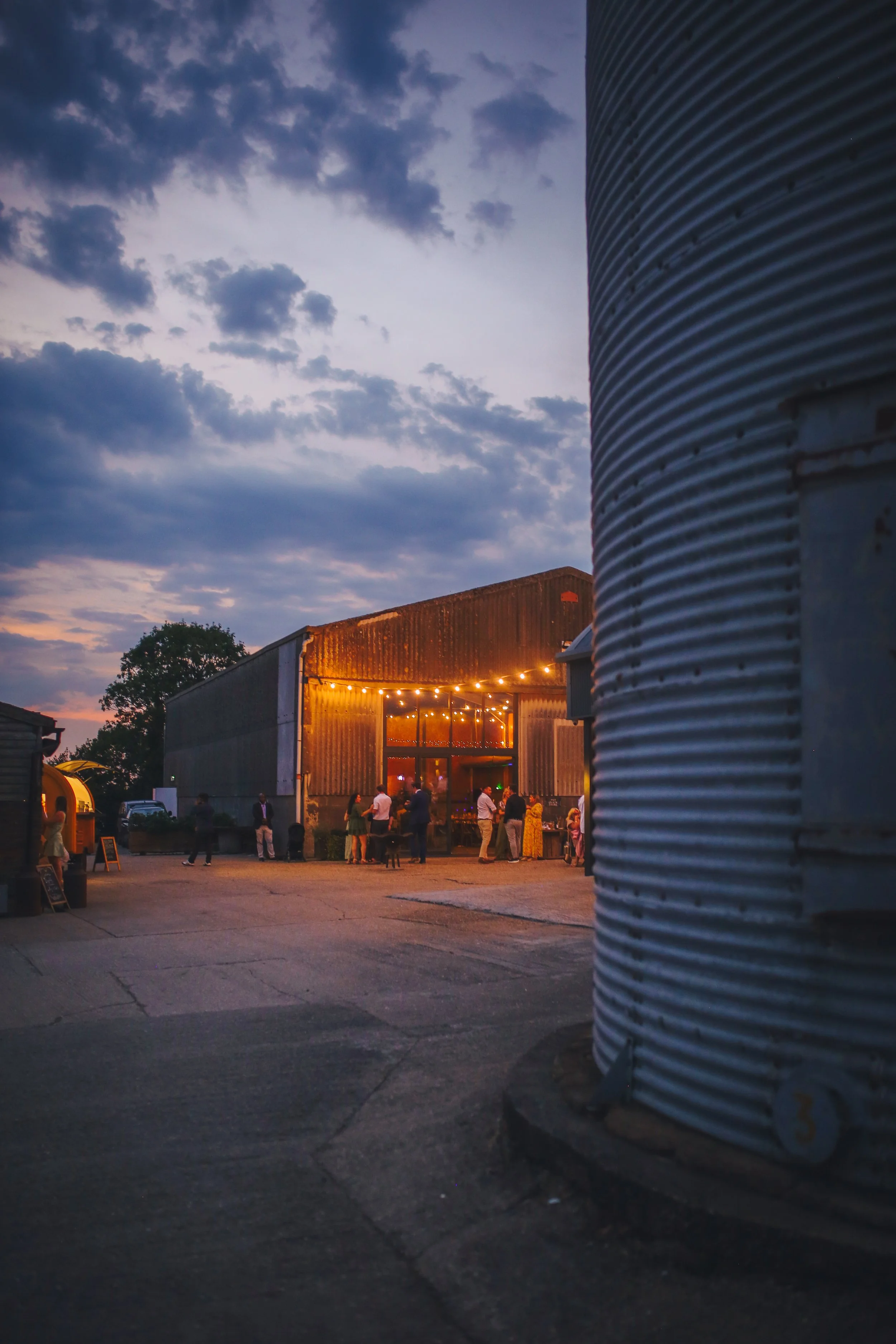 Outdoor gathering at dusk in front of a rustic, industrial-style building with string lights and people socializing.