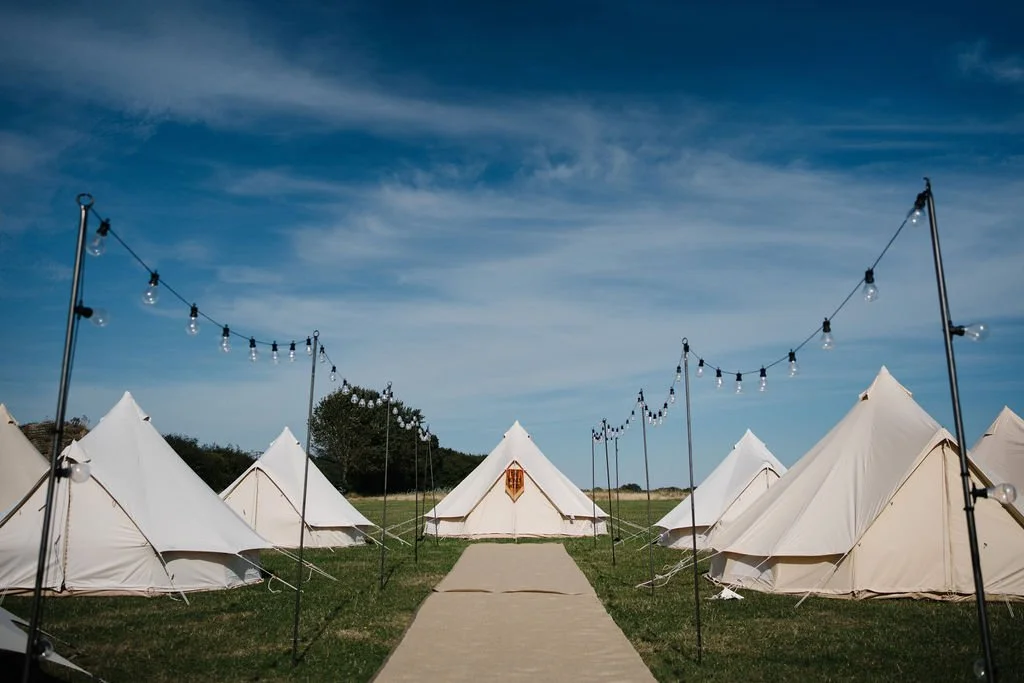 A camp with rows of white tents on a grassy field, a concrete pathway in the middle, and string lights hanging overhead on poles under a partly cloudy blue sky.