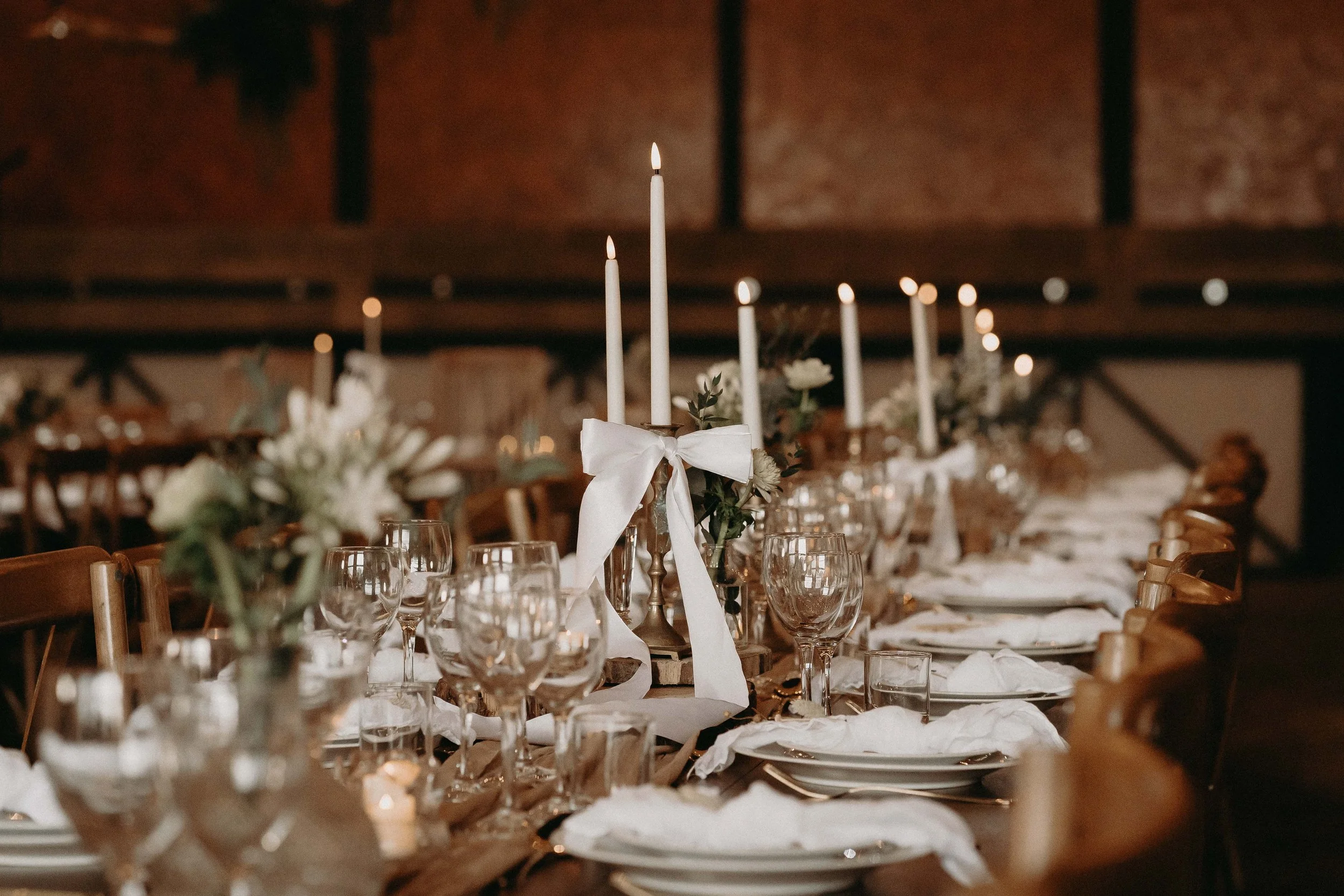 Long dining table decorated with white candles, flowers, glassware, plates, and napkins, set for a formal event in a rustic venue.