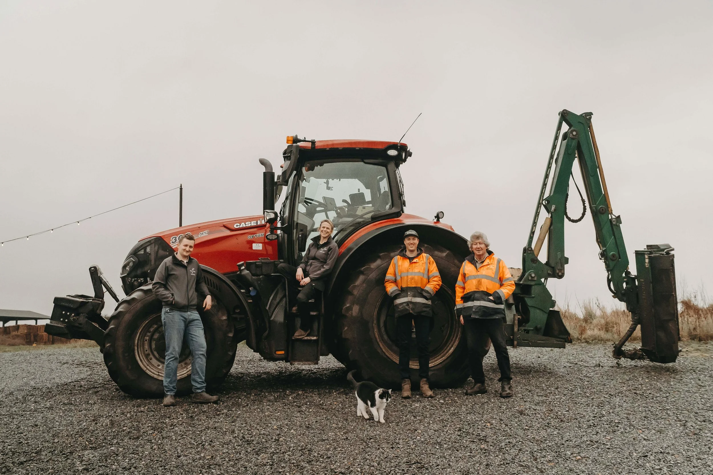 Four people and a cat standing in front of a large red tractor on a gravel surface, with overcast sky in the background.