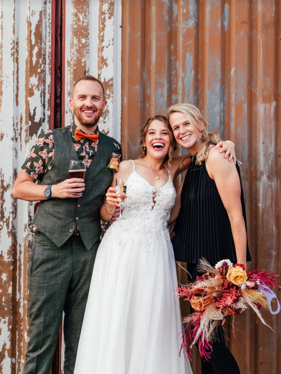 A group of three friends celebrating at a wedding, with one woman in a white wedding dress holding a glass of champagne, a man in a plaid vest and floral shirt holding a drink, and another woman in a black dress holding a bouquet of flowers, all smiling and standing in front of a rustic, weathered metal wall.