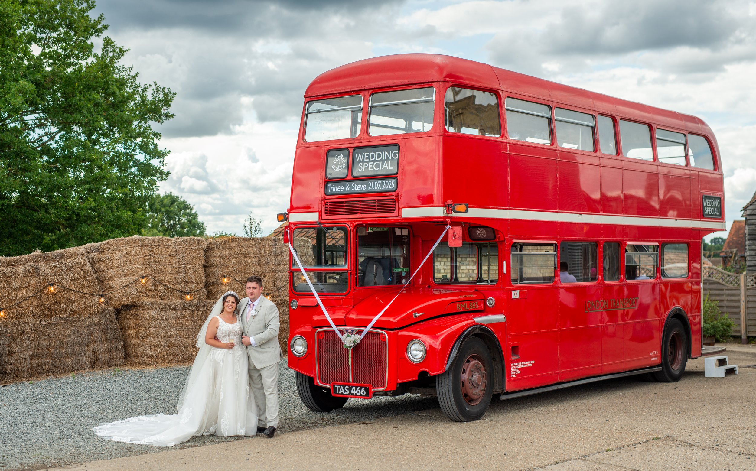 Couple in wedding attire standing next to a red vintage double-decker bus decorated with wedding signs, outdoors on a gravel area with hay bales, cloudy sky, and trees in the background.