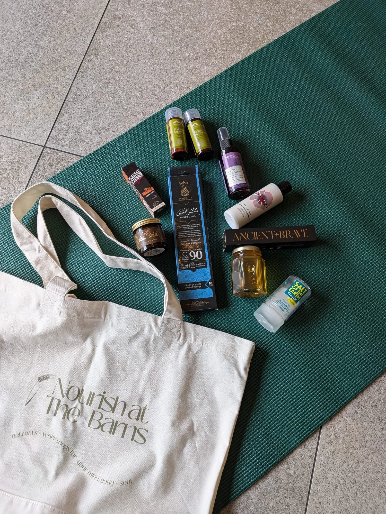 Collection of wellness and personal care products on a green yoga mat, including essential oils, herbal remedies, and skincare items, with a beige tote bag reading 'Nourish at The Barns'.