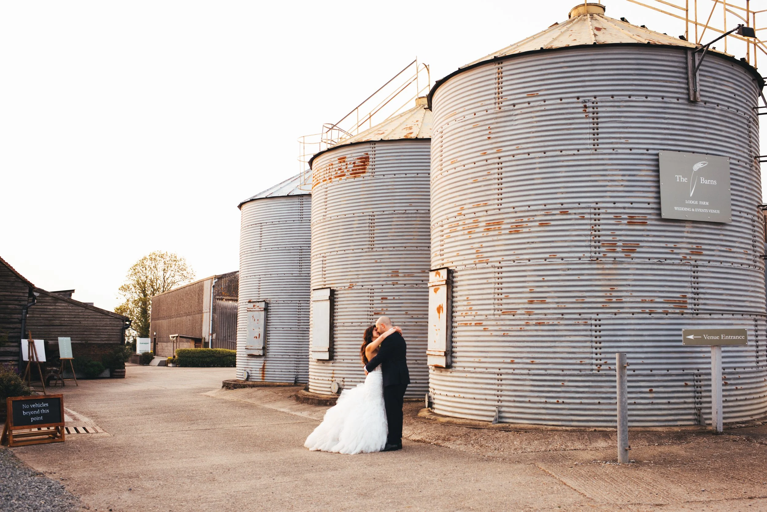 A bride and groom embrace and kiss in front of three large, rusty grain silos outside a venue called The Barns during sunset.
