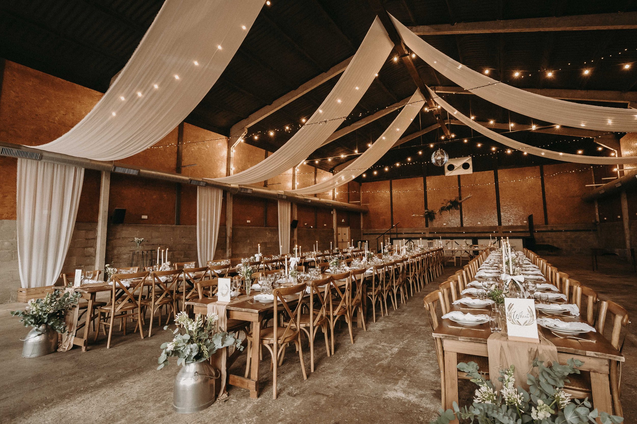 A large indoor event space with wooden tables and chairs arranged for a formal gathering or wedding reception. The ceiling is decorated with white fabric drapes and string lights, creating a warm and festive atmosphere. The tables are set with white plates, glasses, candles, and floral centerpieces.