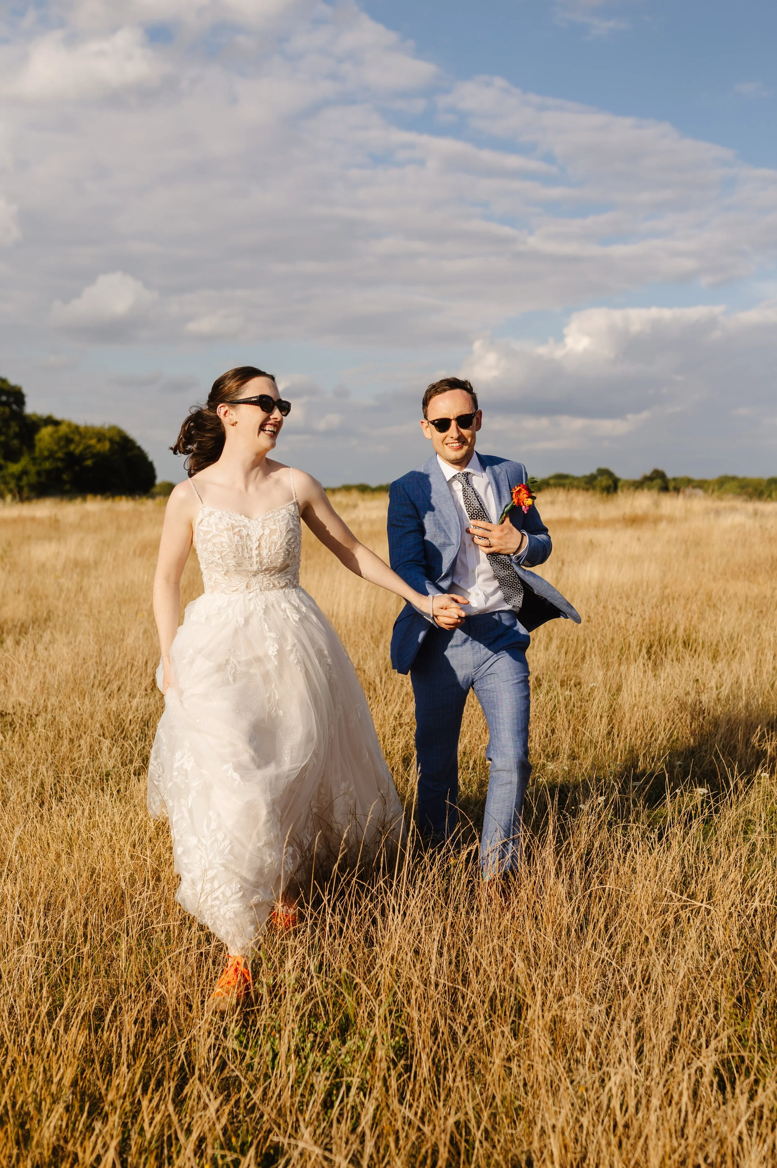 A smiling newlywed couple walking through a grassy field on a sunny day, with the bride in a white wedding gown and the groom in a blue suit.