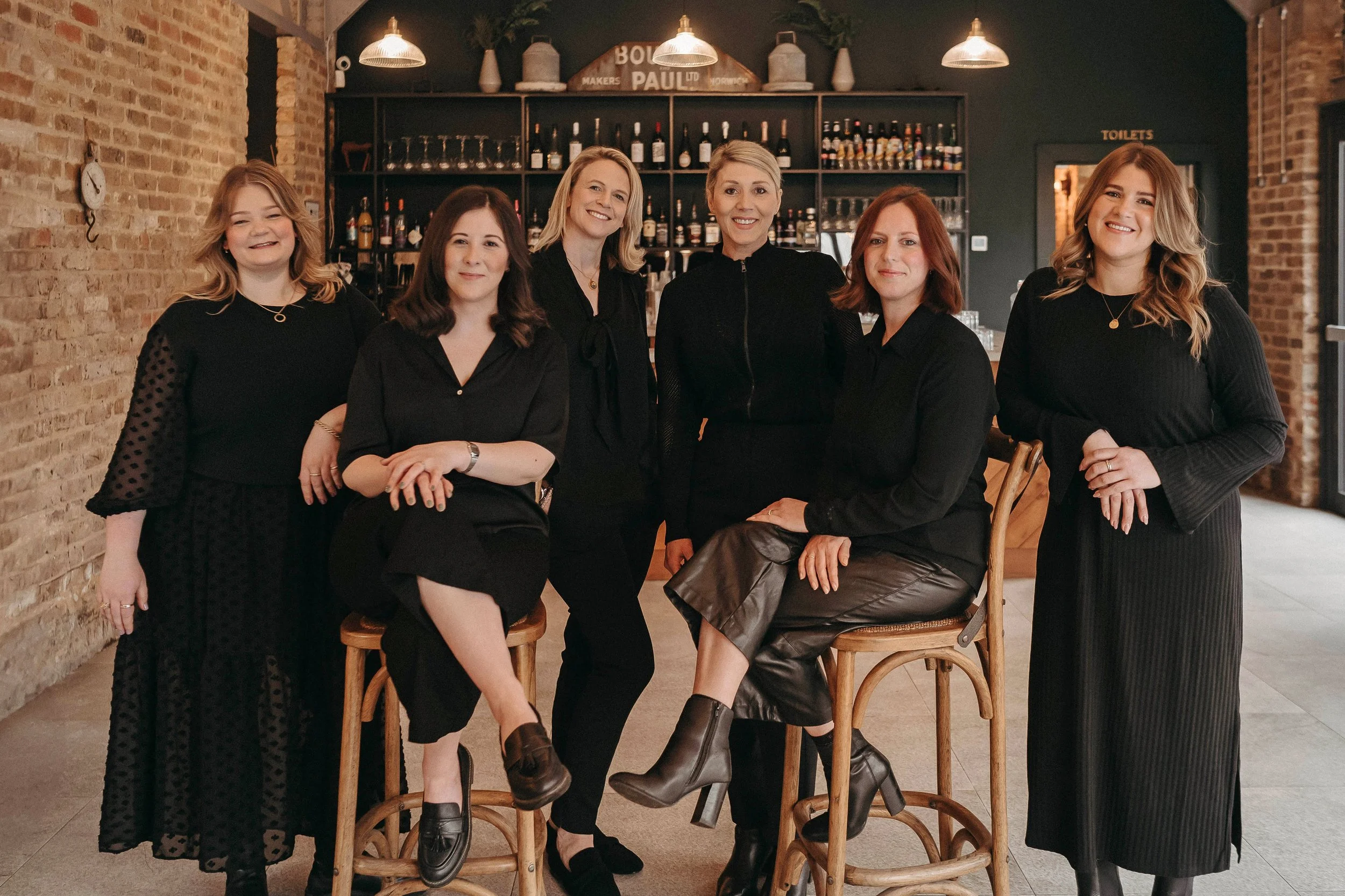 Six women dressed in black, posing in a restaurant or bar with a brick wall and shelves of wine bottles in the background.