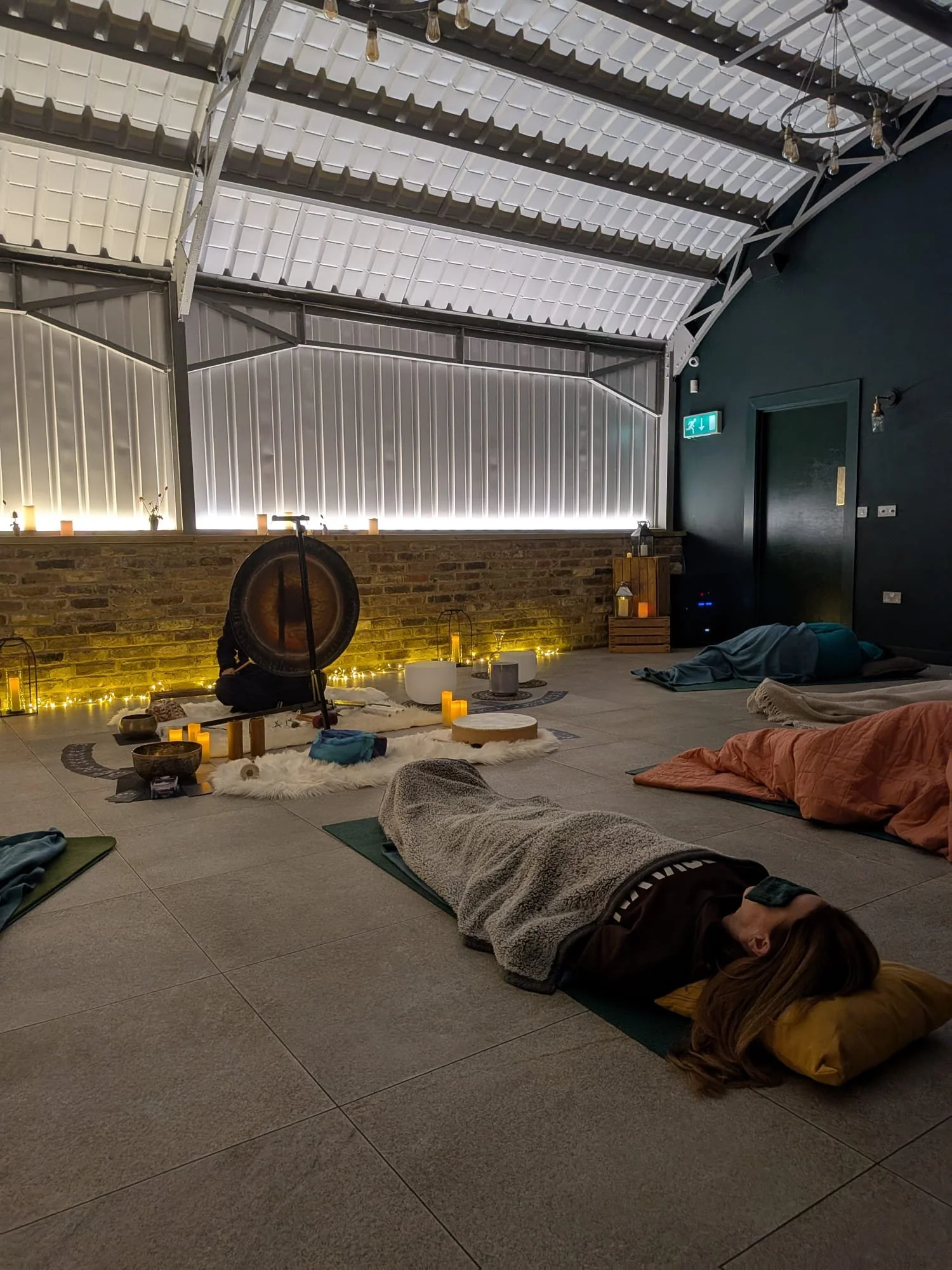 People lying on mats and blankets in a dimly lit room with candles, velvet cushions, and a gong in the background, suggestive of a meditation or yoga session.