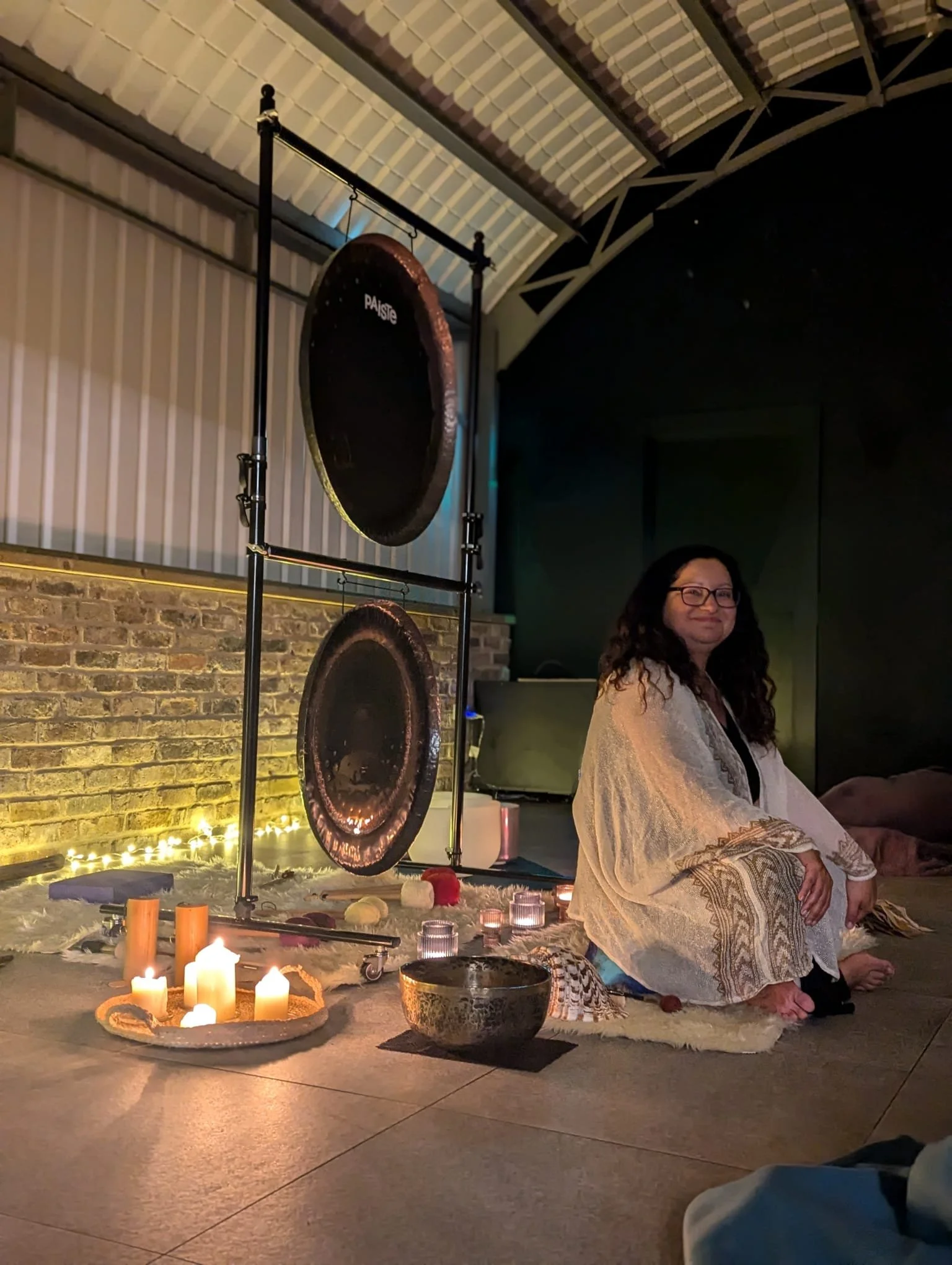 A woman sitting cross-legged on the floor in front of a large gong with candles and decorative objects around her, in a dimly lit room.