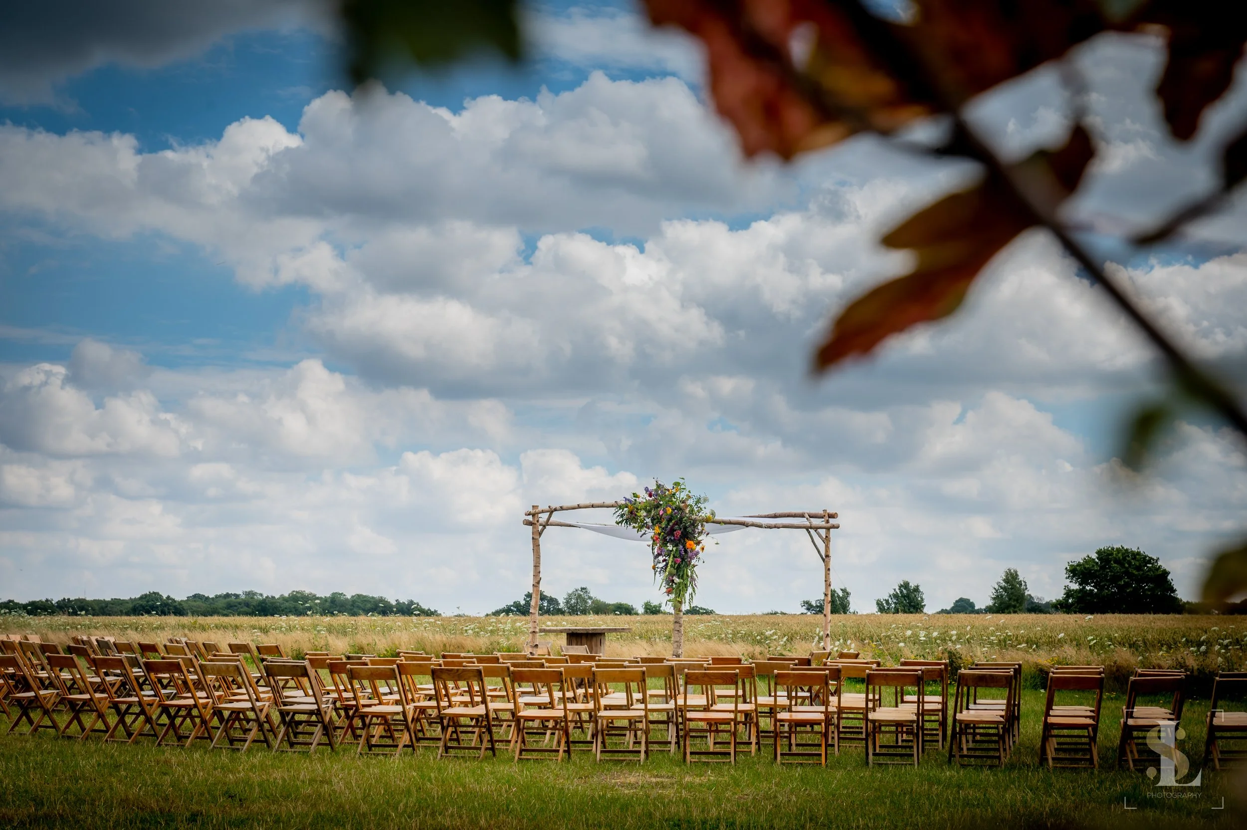 Outdoor wedding ceremony setup with rows of wooden chairs facing an arch decorated with colorful flowers, on a grassy field under a partly cloudy sky.