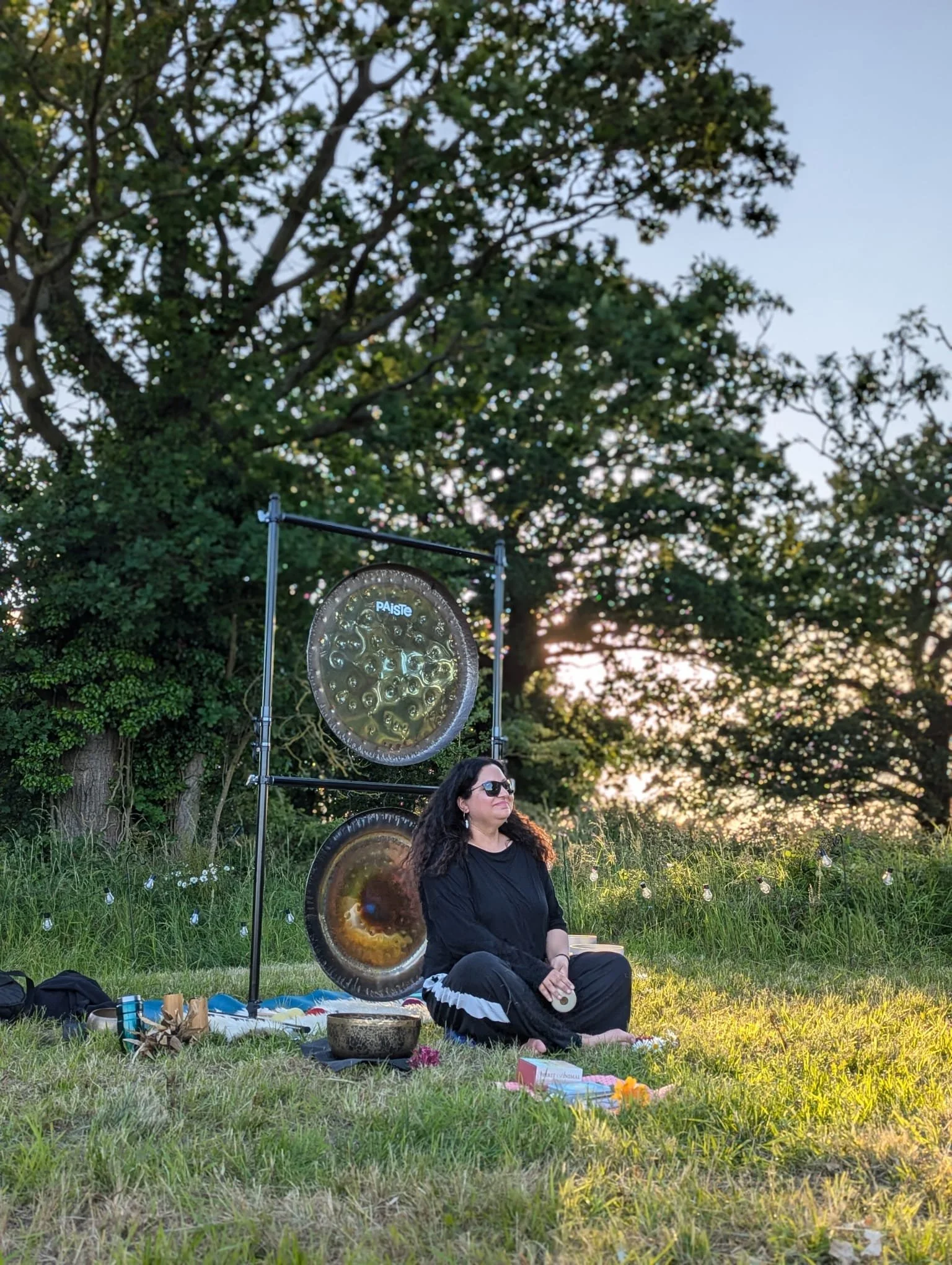A woman with curly hair and sunglasses sits cross-legged on the grass at sunset, next to large hanging gongs and a singing bowl, with trees and a sky in the background.