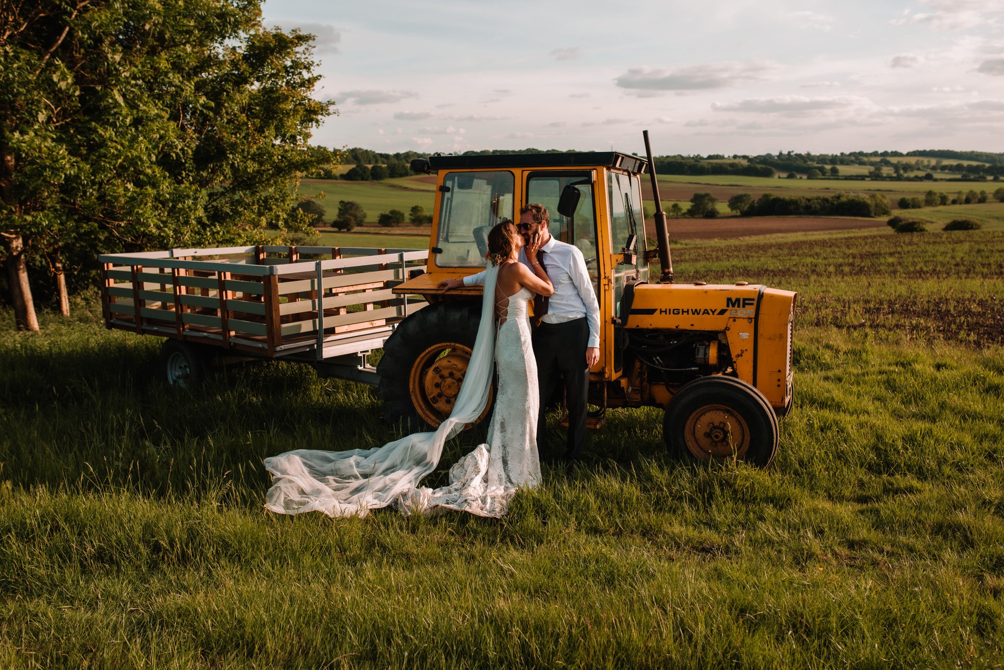 A bride and groom kissing next to a yellow tractor in a grassy field during sunset, with a tree and open farmland in the background.
