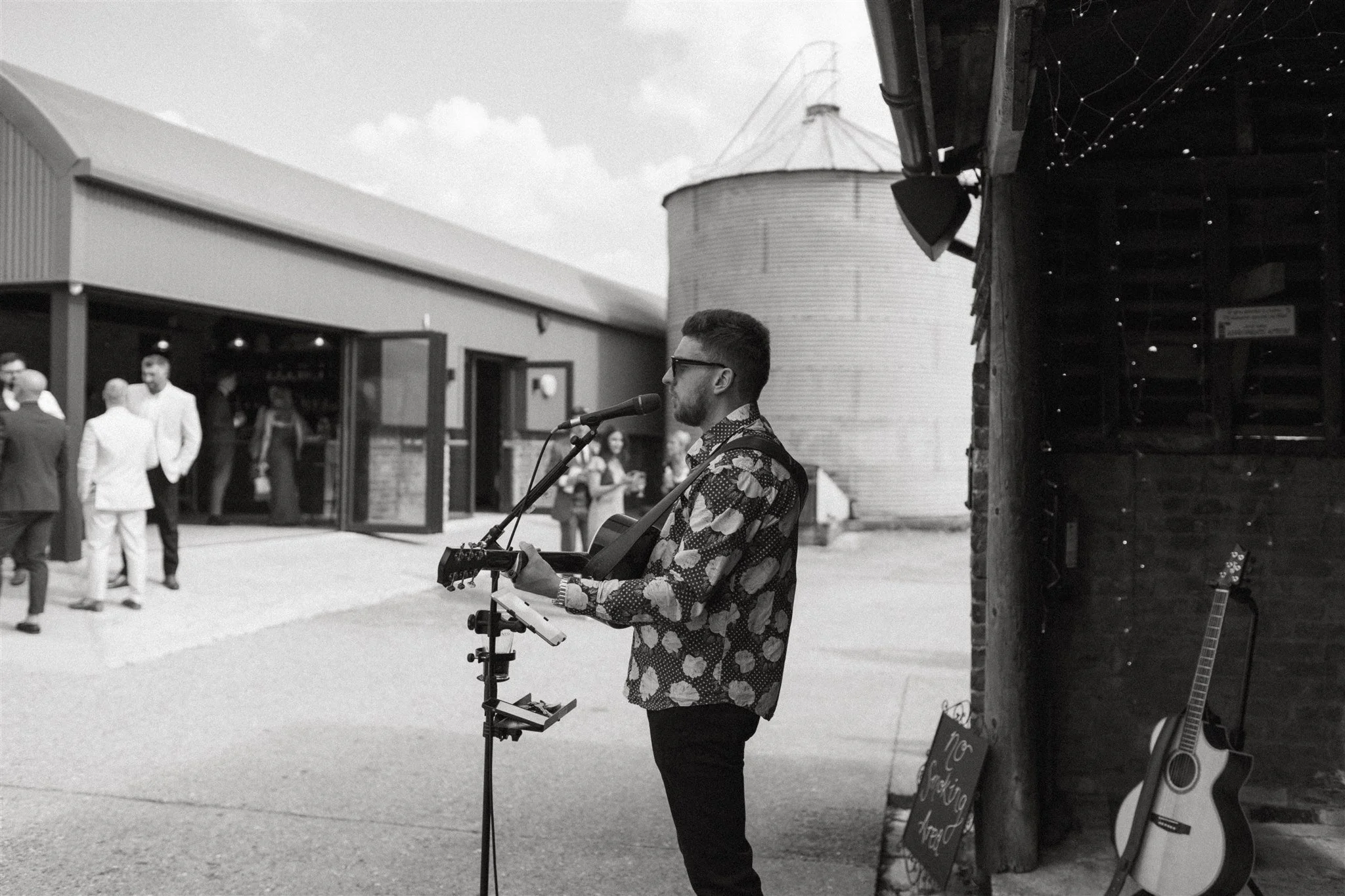 A young man with sunglasses, wearing a floral shirt, playing an acoustic guitar and singing into a microphone outside a rustic venue. There are people in formal attire in the background, some chatting, a chalkboard sign, and another guitar leaning against a brick wall next to a small stand.