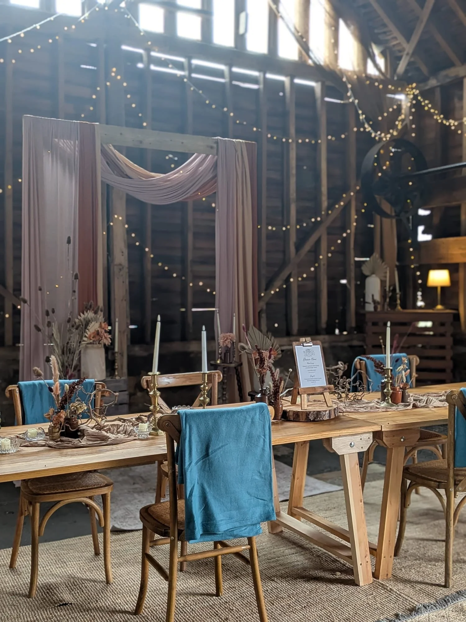 A rustic wedding setup inside a barn with a wooden table decorated with dried flowers, candles, and a frame holding a menu, surrounded by mismatched chairs with blue cloth draped over some, and string lights hanging from the barn walls and ceiling.