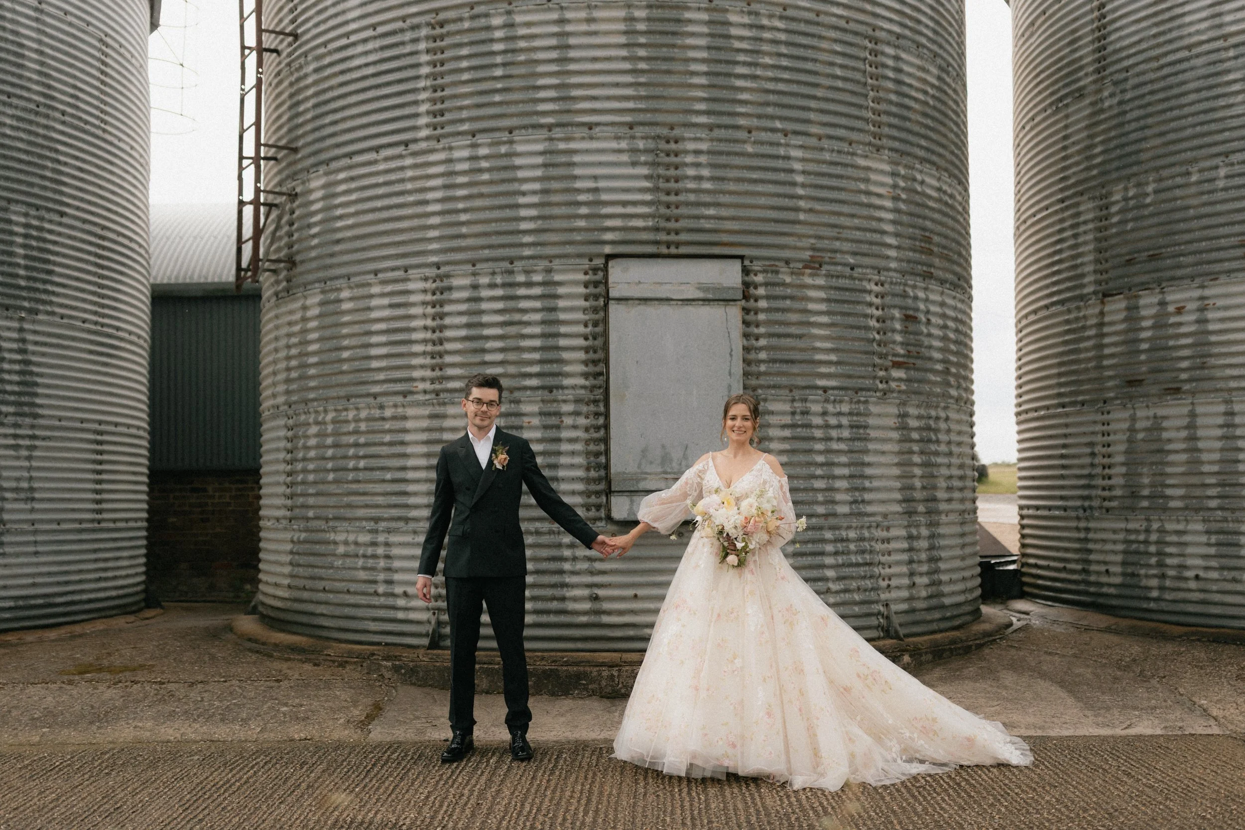 A bride and groom holding hands, standing in front of large metal silos, outdoors on a cloudy day. The bride is wearing a white wedding gown holding a bouquet of flowers, and the groom is wearing a black tuxedo with a boutonniere.