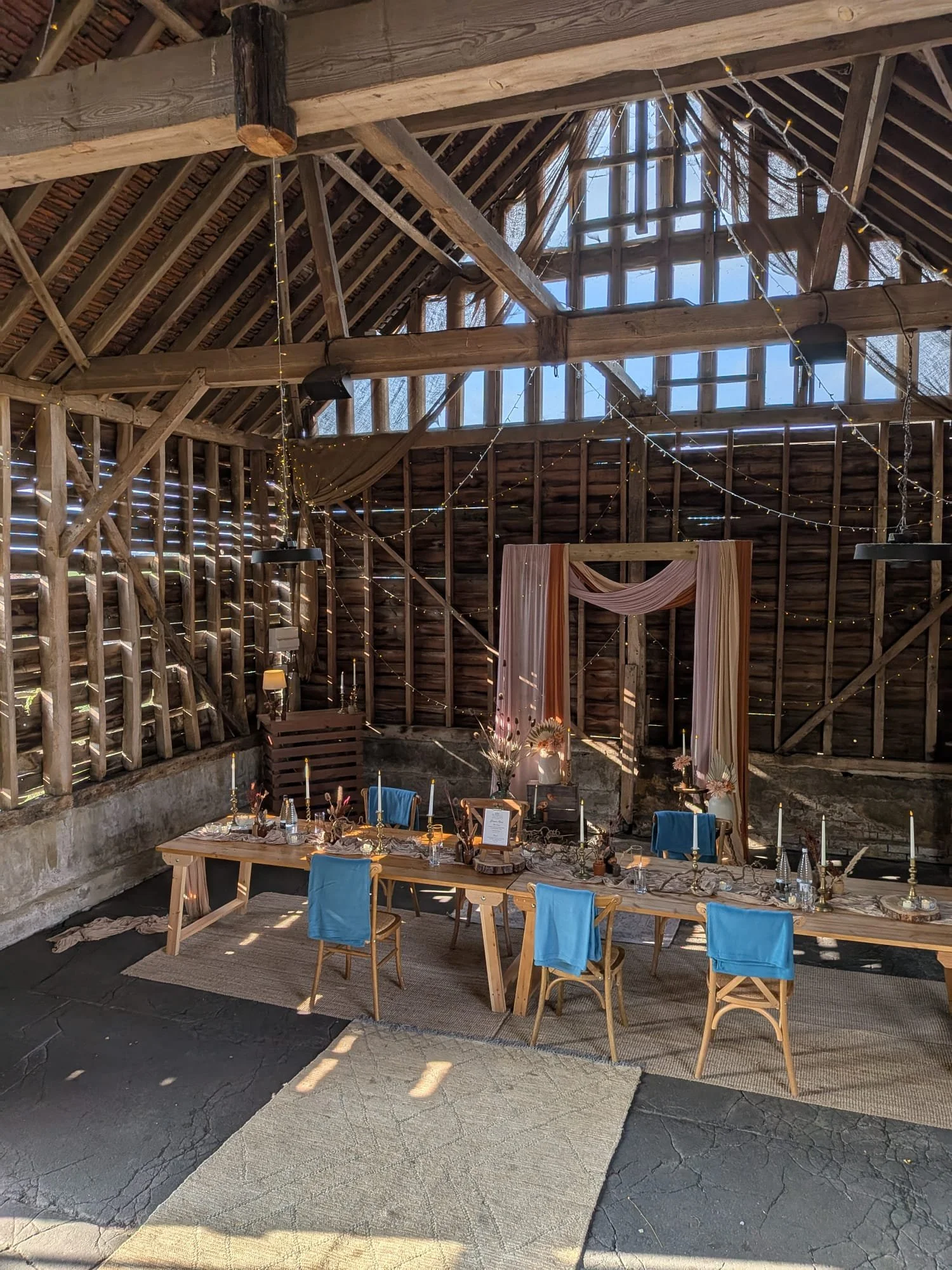 A rustic barn wedding setup with wooden chairs arranged in rows on either side of a central aisle lined with bouquets. The barn has wooden walls and a high, exposed beam ceiling decorated with string lights, creating a warm and festive atmosphere.