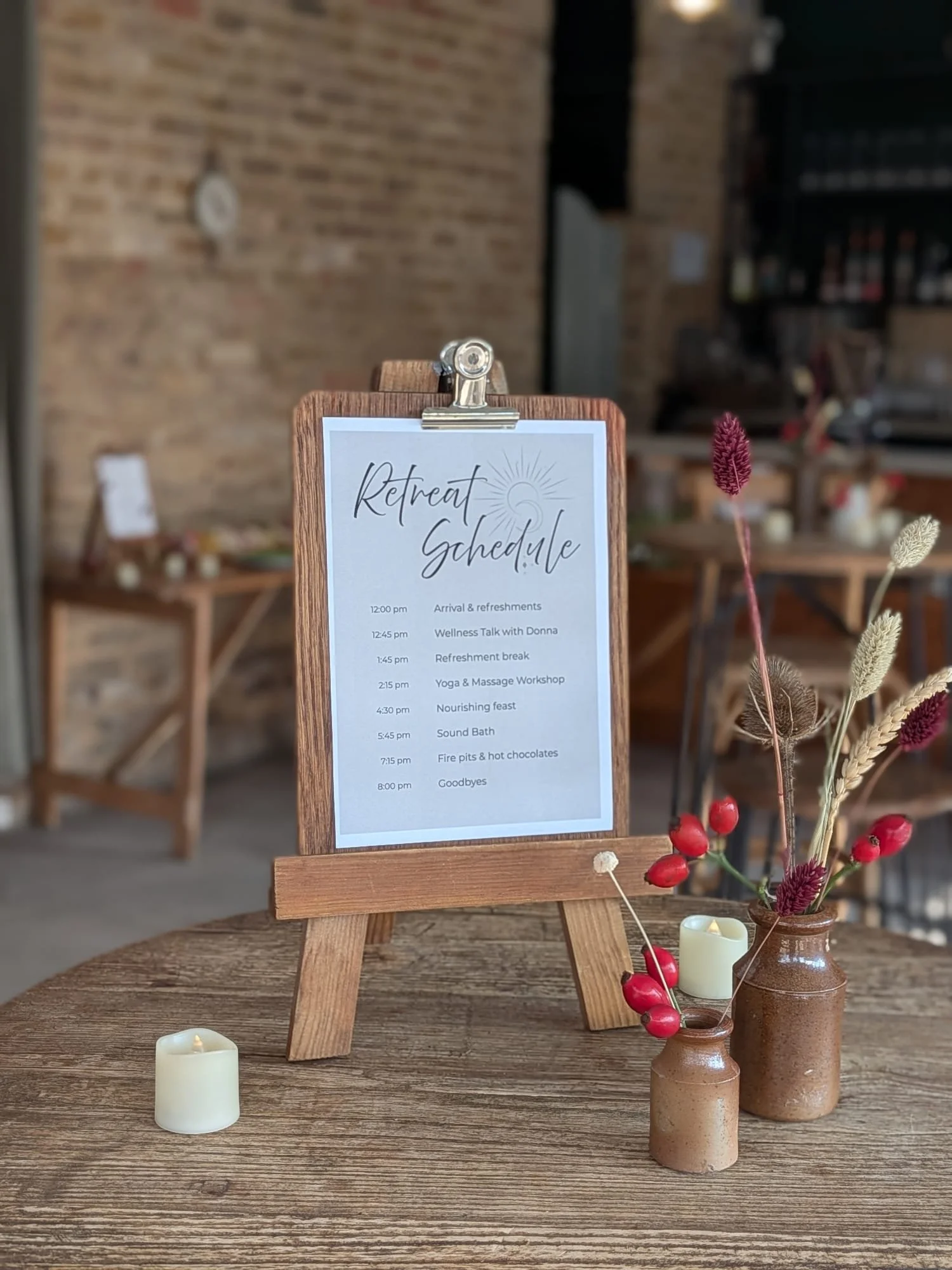 Wooden table holding a small framed schedule titled 'Retreat Schedule' with a list of activities and times, next to two brown vases with dried flowers and red berries, and a small candle.