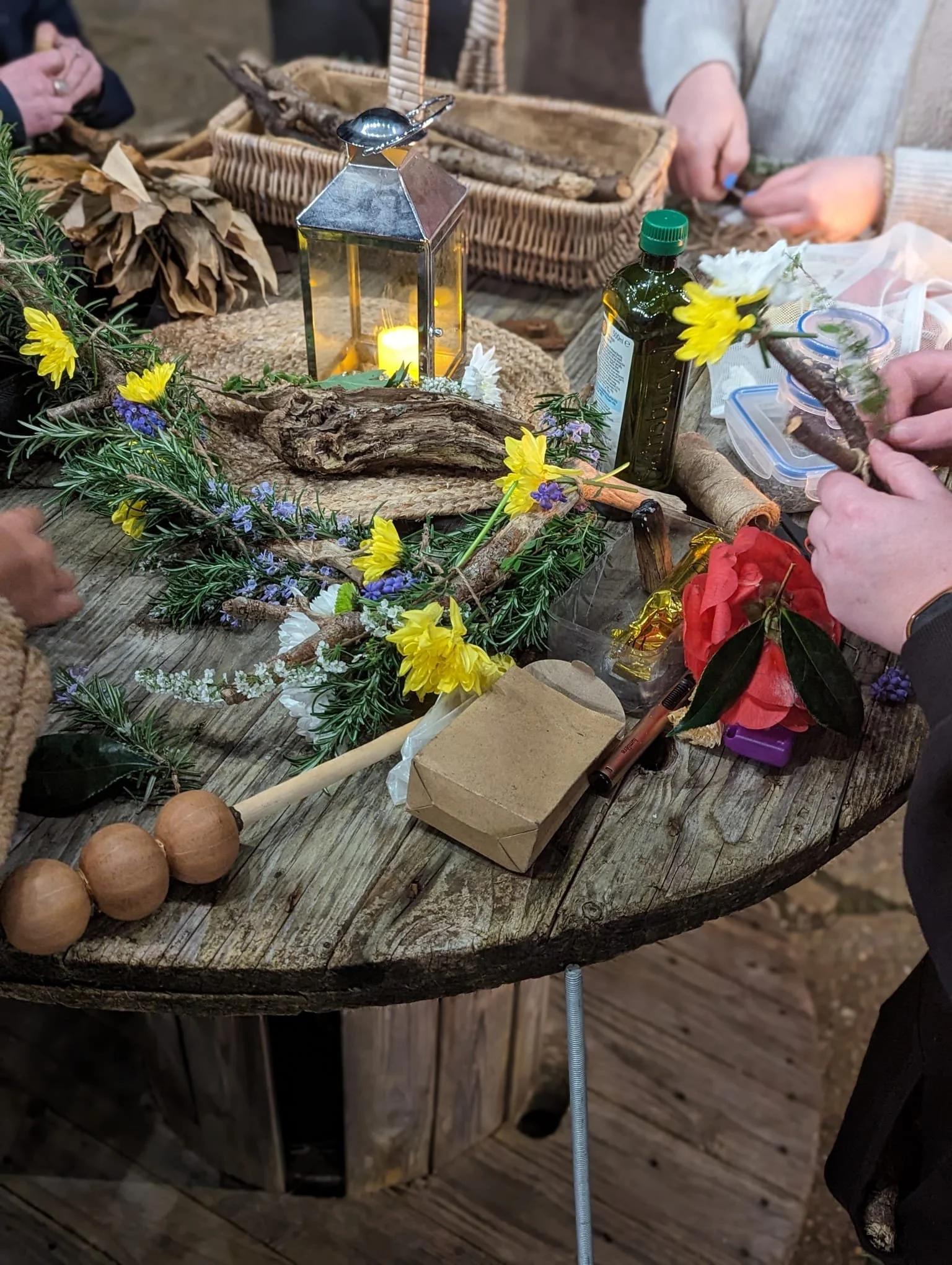 People creating floral arrangements with flowers and greenery on a rustic wooden table decorated with a lantern, driftwood, and craft supplies.