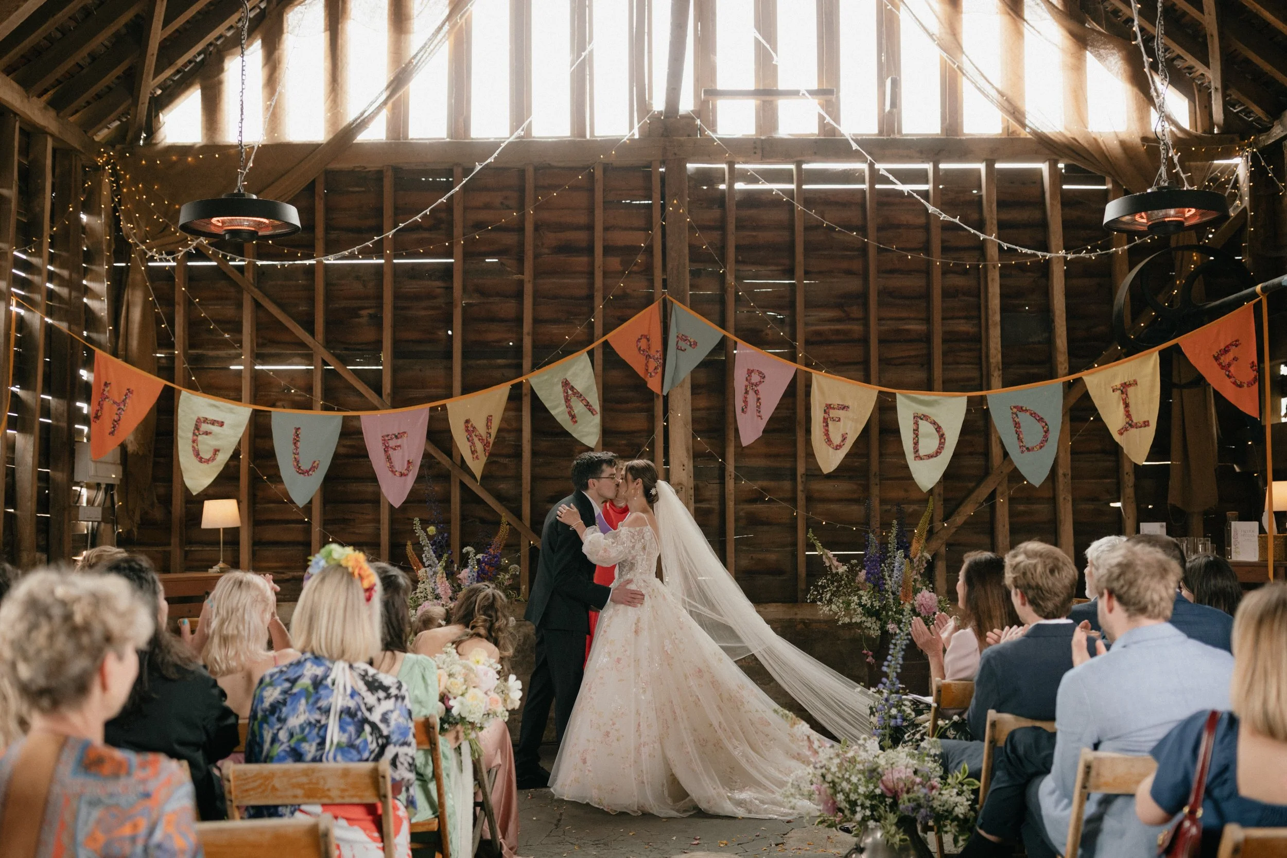 A bride and groom kiss during their wedding ceremony in a rustic barn decorated with string lights and a colorful banner reading 'Loved and Married.' Guests sit in wooden chairs watching and clapping.