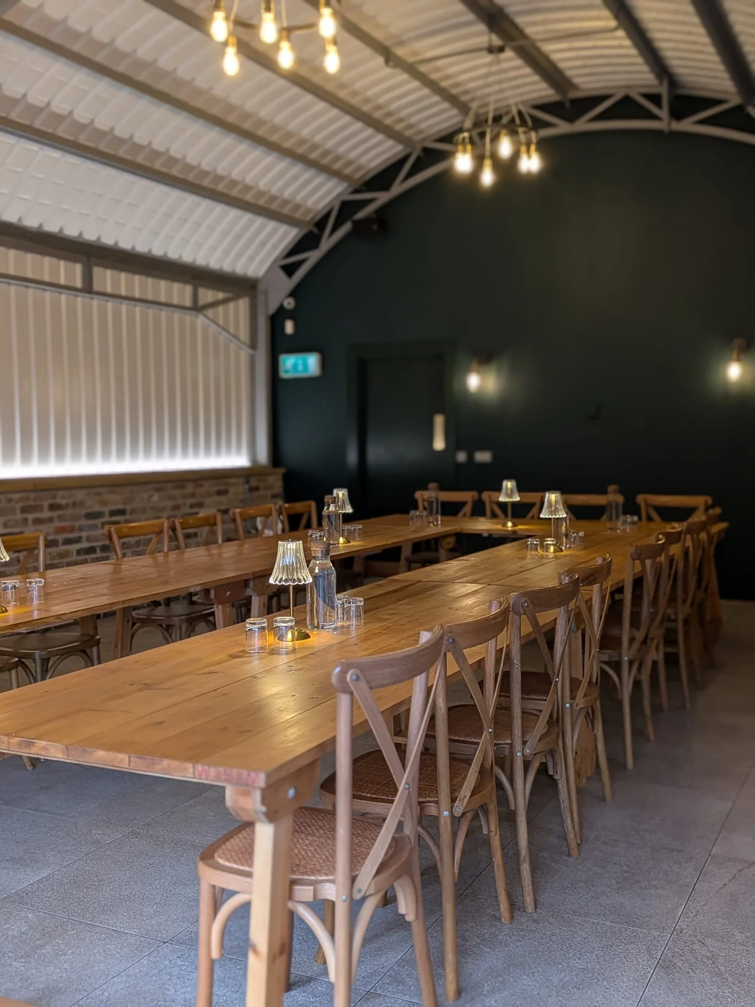 Interior of a modern bar with a curved metal roof, wooden furniture, a dark green wall, and a fully stocked bar counter.