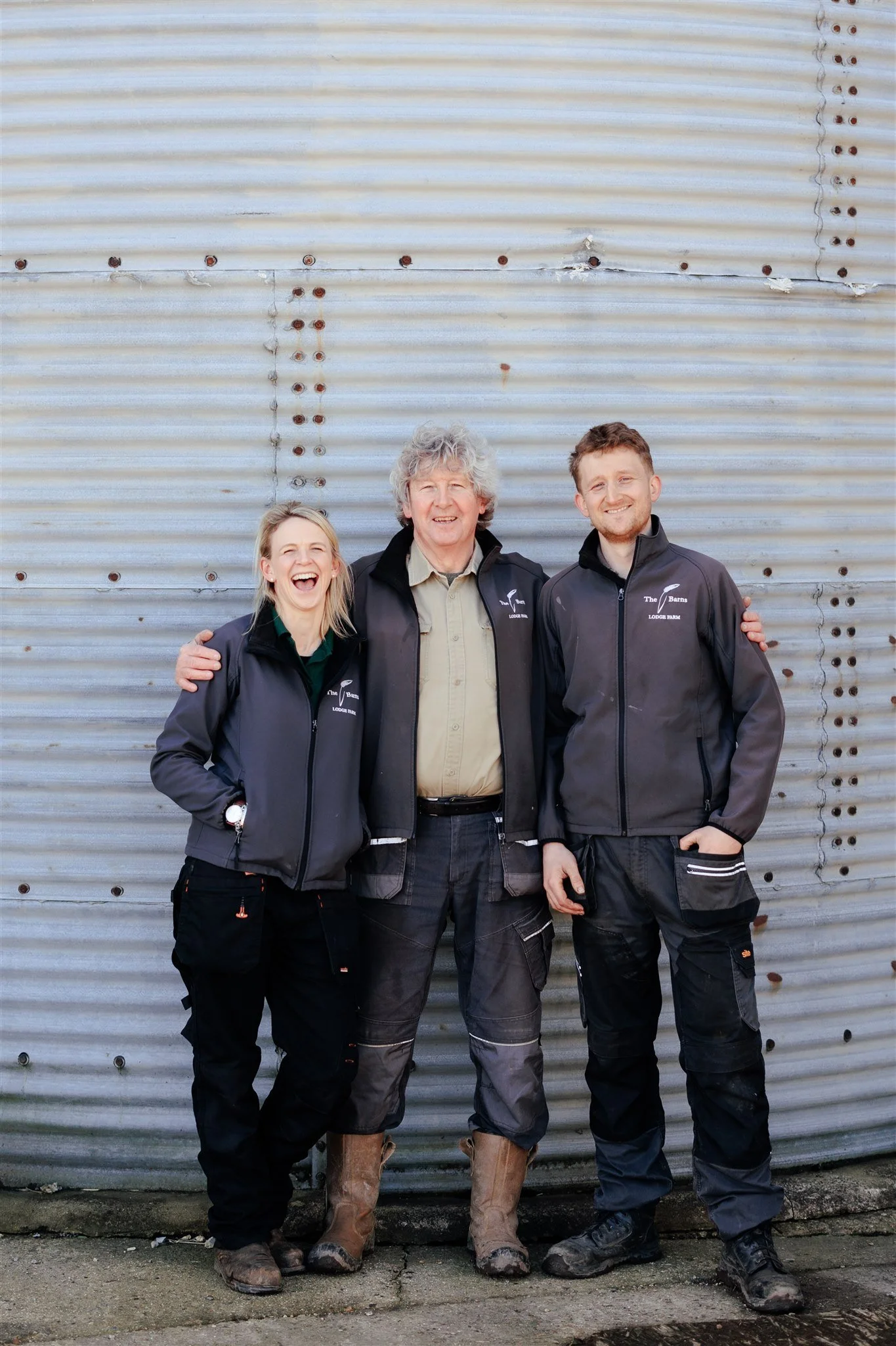 Three people, two men and one woman, standing together outdoors against a corrugated metal wall. They are wearing matching dark jackets with a logo that says "The Barns Lodge Farm". They are smiling and appear happy.