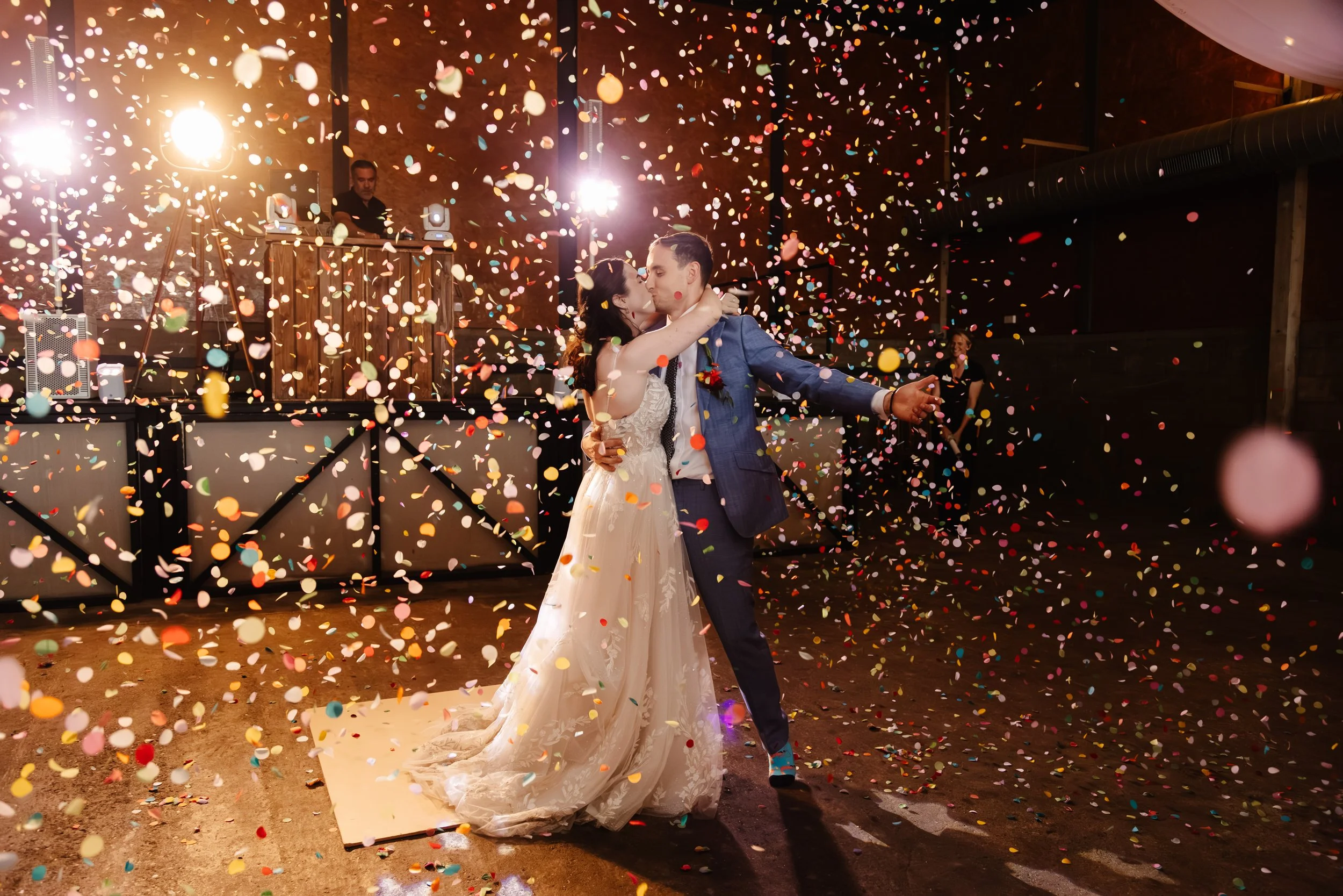A newlywed couple sharing a kiss on the dance floor during their wedding reception, surrounded by colorful confetti and illuminated by bright lights, with a DJ playing music in the background.