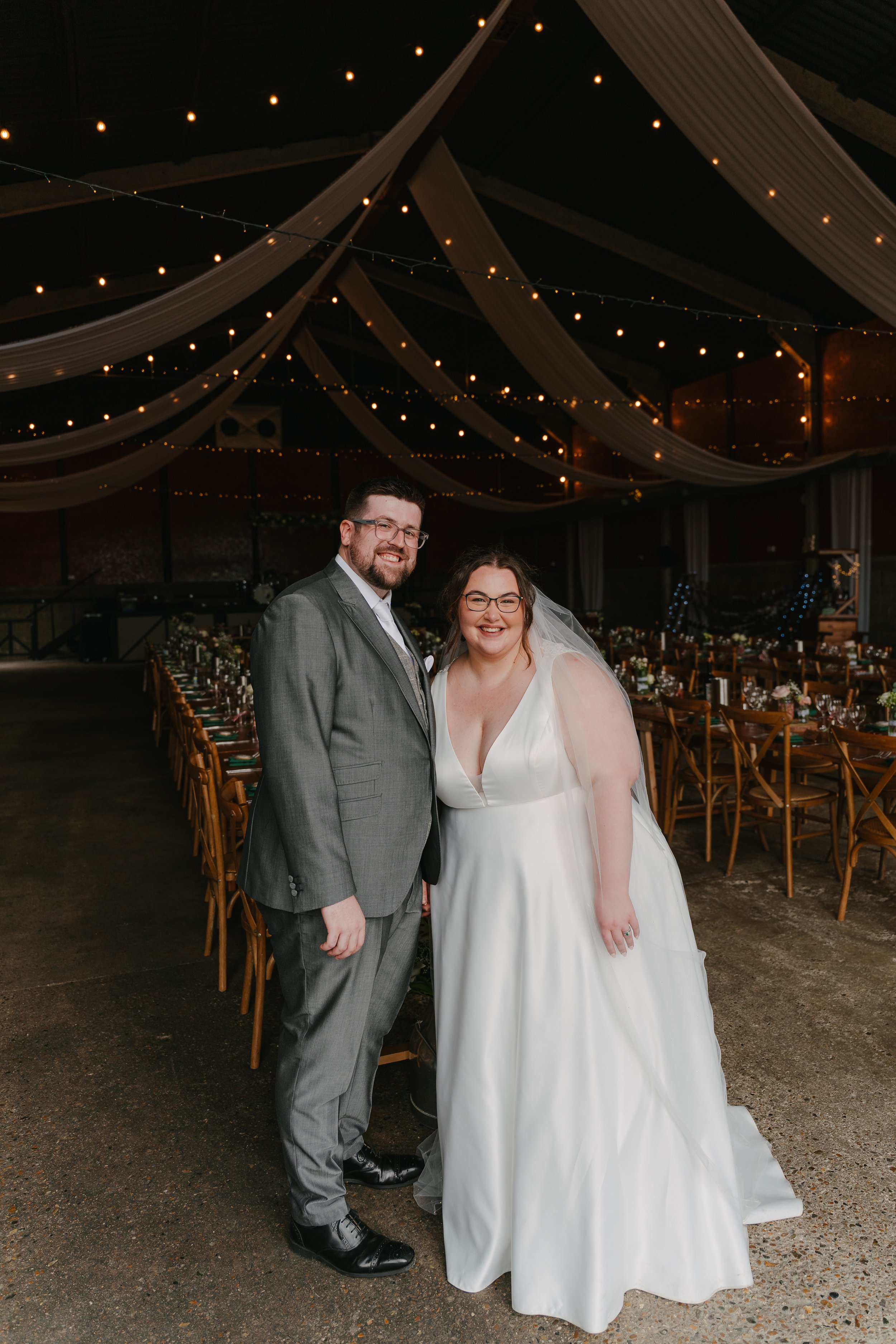 A bride and groom standing together at their wedding reception. The bride is wearing a white wedding dress and a veil, and the groom is dressed in a grey suit. The reception venue has draped fabric and string lights hanging from the ceiling, with ban
