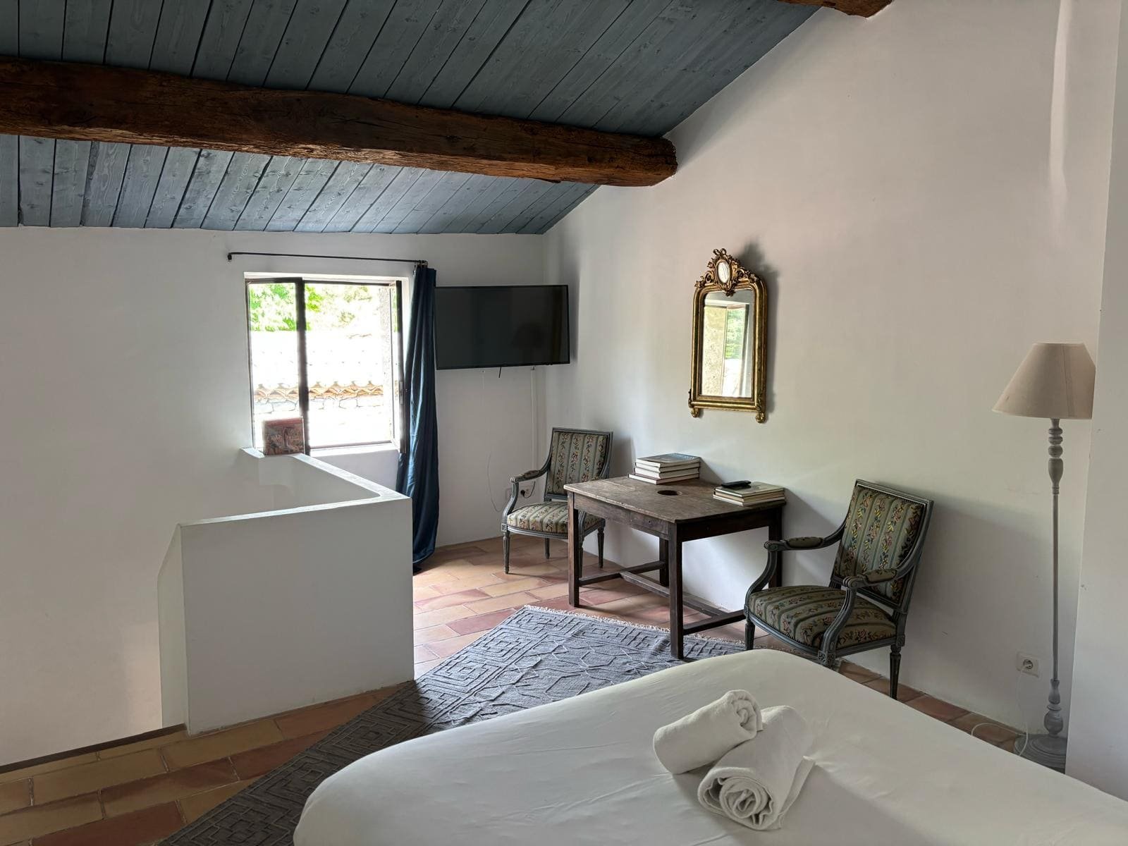 Chambre avec lit, deux fauteuils, un bureau avec livres, miroir doré, télévision, fenêtre avec rideaux, lampe sur pied, planche de bois au plafond, sol en tuiles.