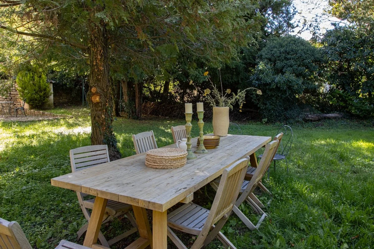 Table en bois avec des chaises en bois, décorée de bougies, un vase avec des fleurs et des assiettes, dans un jardin verdoyant avec des arbres.