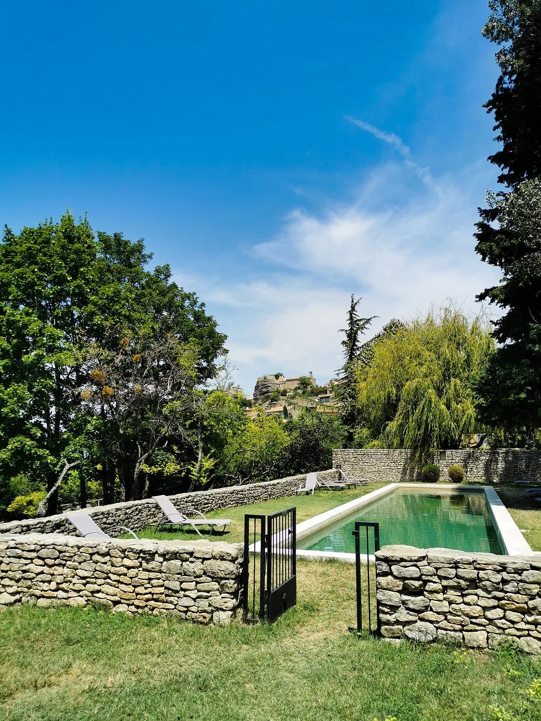 Piscine entourée d'une clôture en pierre, avec des chaises longues autour, des arbres verts et un ciel bleu en arrière-plan.