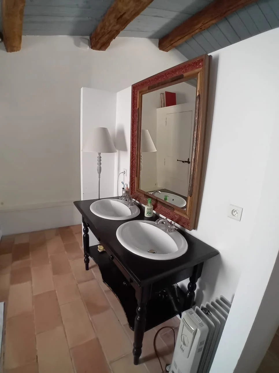 Un grand miroir avec un cadre en bois rouge-dessus arrondi, deux vasques blanches sur un meuble noir avec deux lampes de chaque côté. La pièce a un plafond en bois et un sol en carrelage beige.