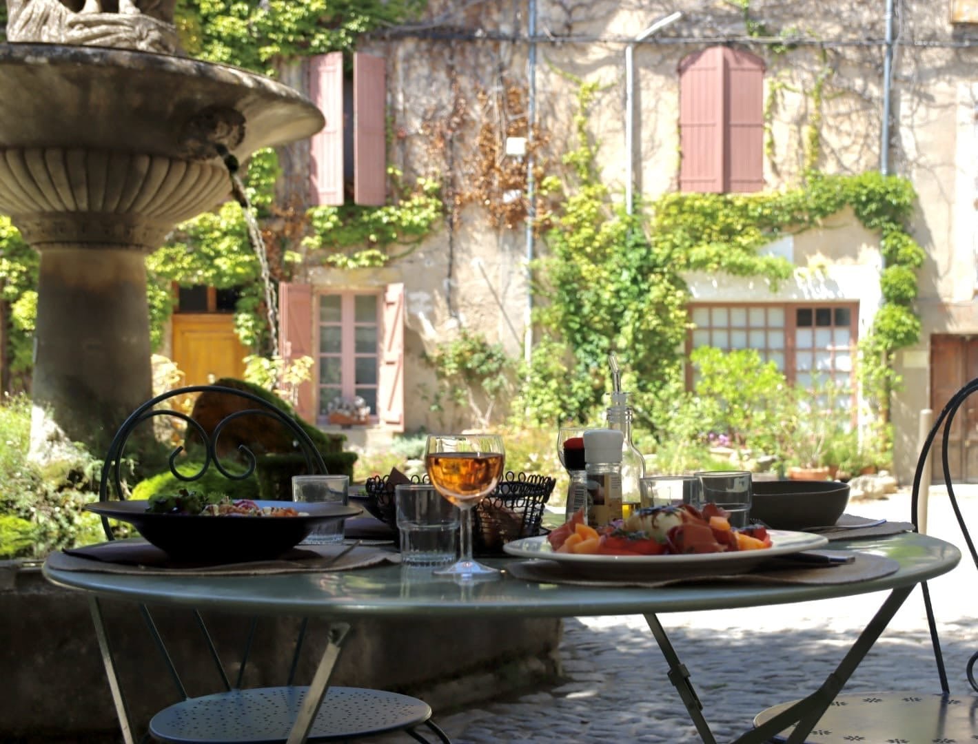 Repas en plein air avec vue sur une fontaine, assiettes de nourriture, verres et boissons sur une table, dans un jardin avec murs en pierre, fenêtres à volets et végétation luxuriante.