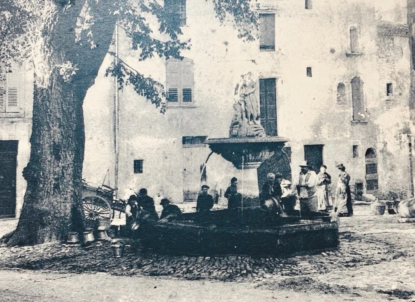 An old photograph showing people around a fountain in a building courtyard with trees and old buildings in the background.